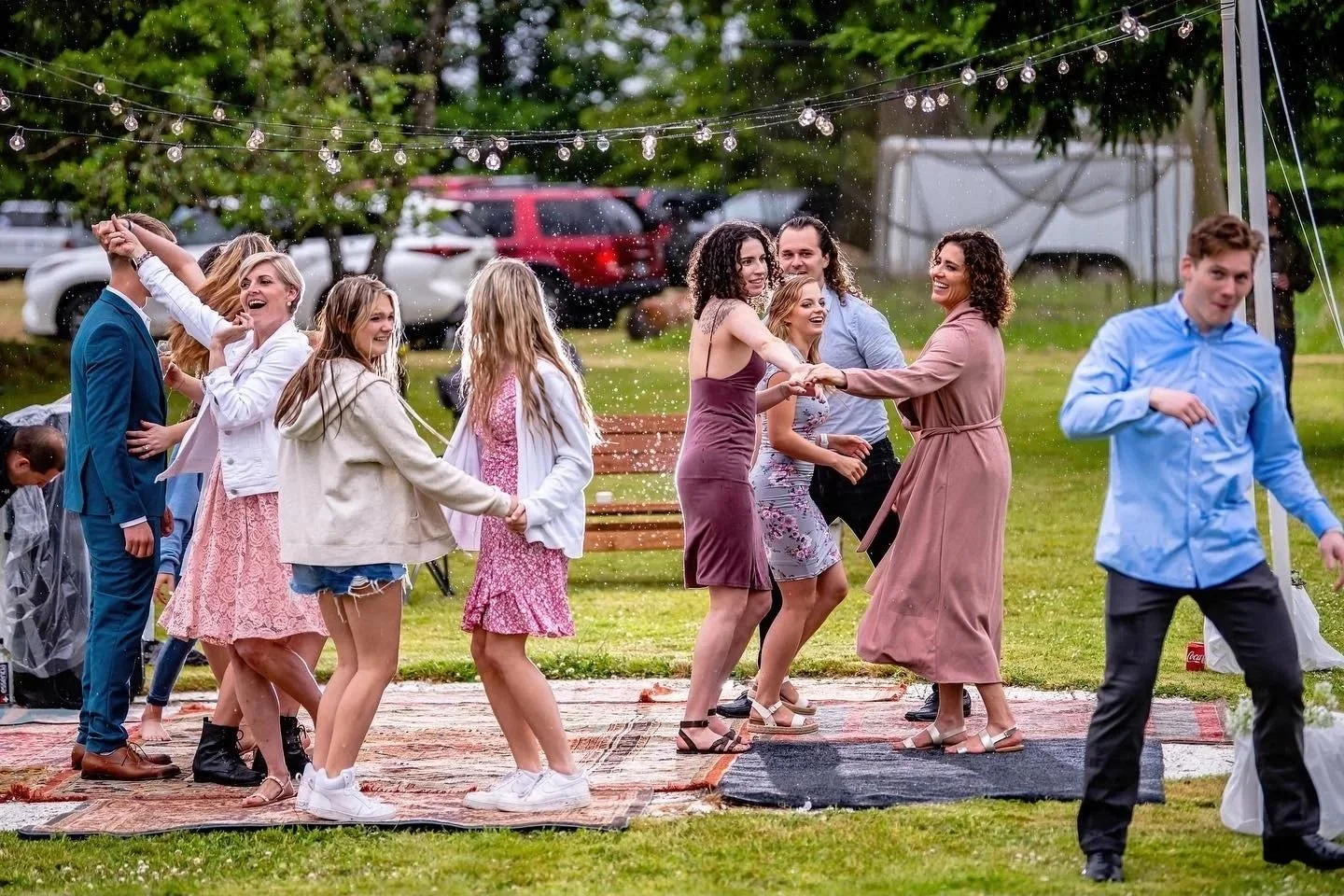 People dancing and celebrating outdoors under string lights, with some holding hands and others spinning, on a grassy area with trees and parked cars in the background.