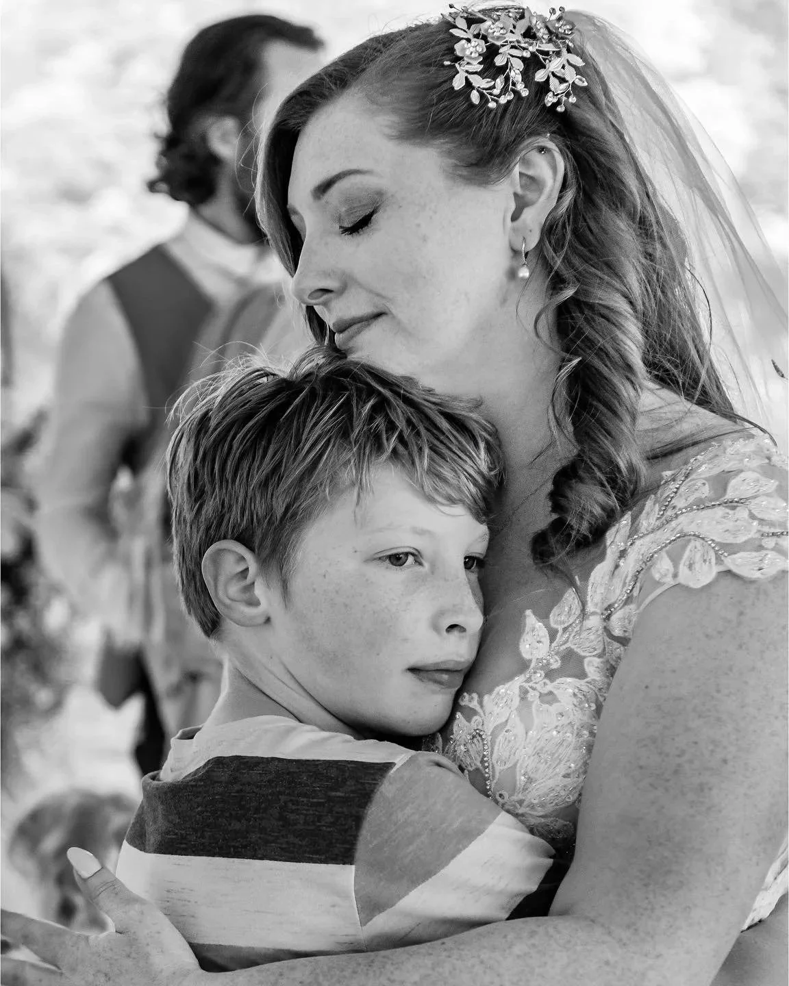 A woman with a floral headpiece and pearl earrings hugging a young boy with freckles, in black and white, at a heartfelt moment.