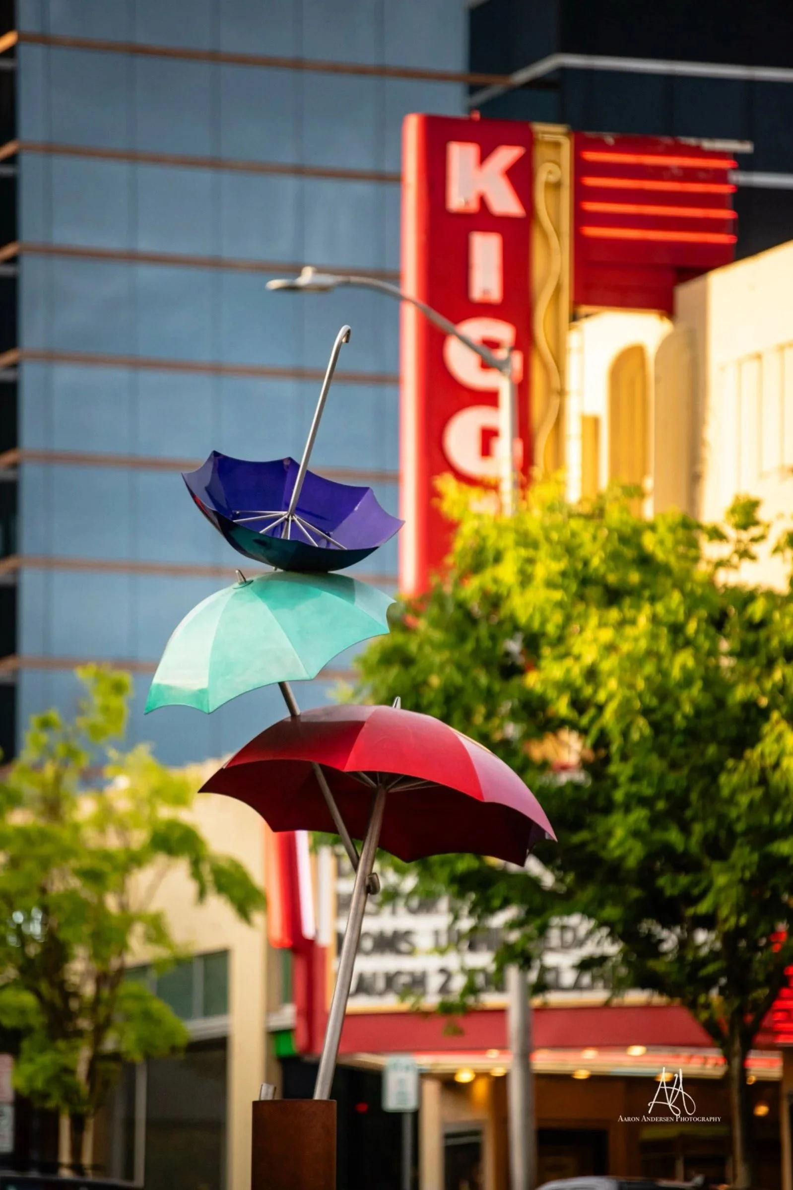 Colorful umbrellas stacked on thin poles in an urban setting, with trees and a building in the background, including a red vertical neon sign.