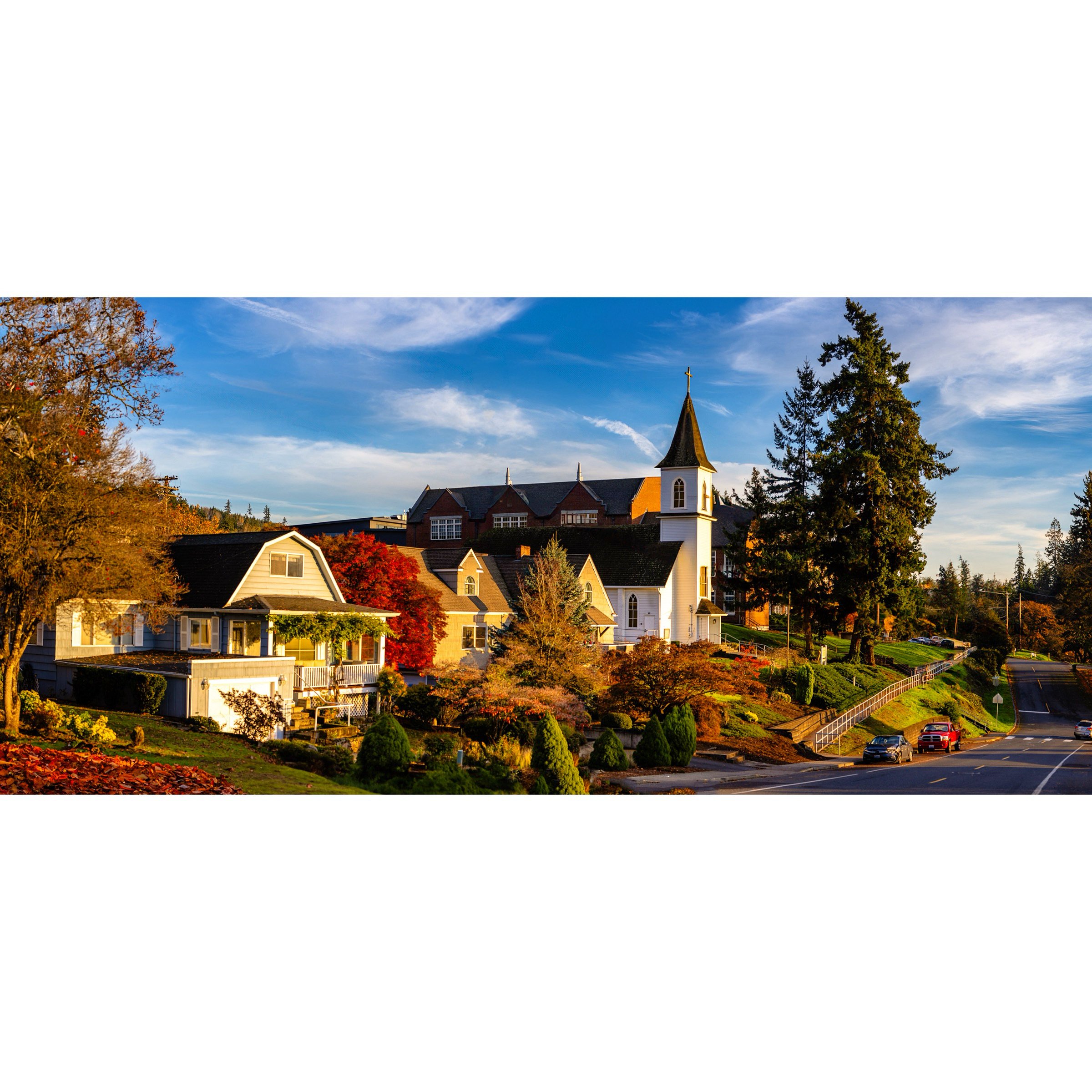 A scenic view of a small town with houses, trees with autumn foliage, and a church with a tall steeple, under a blue sky with white clouds.
