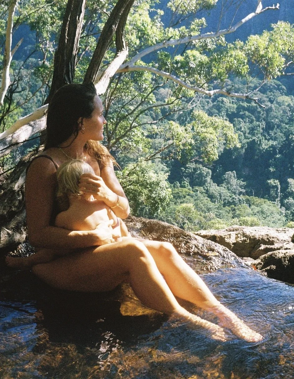 A woman sitting on rocks in a natural stream, holding a child, with trees and greenery in the background.