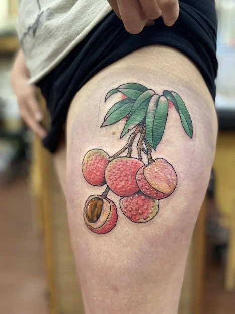 A colorful tattoo of lychee fruits with green leaves on a person's upper thigh.