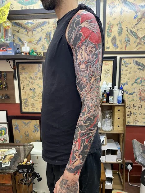 Close-up side view of a man with a colorful full sleeve tattoo on his right arm, standing in a tattoo shop with various tattoo designs and supplies in the background.
