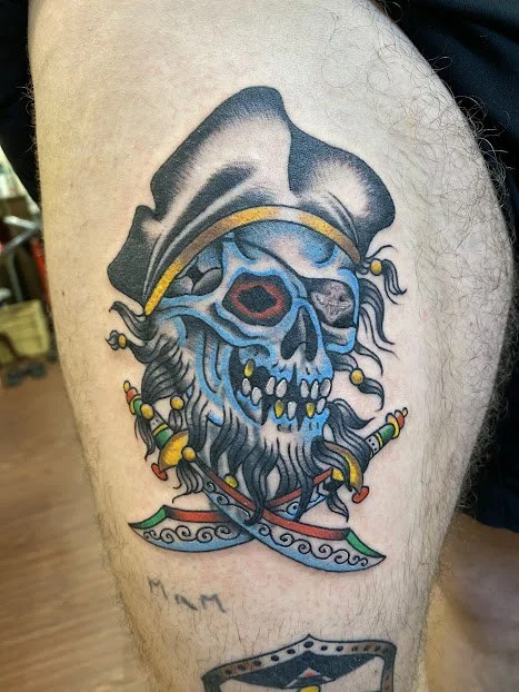 A colorful tattoo of a skull wearing a pirate hat, with a beard and mustache, surrounded by decorative elements.
