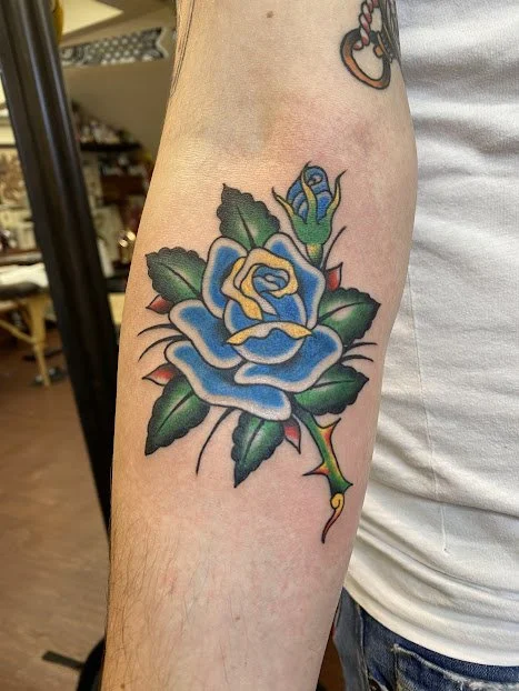 Tattoo of a blue rose with green leaves on a person's forearm.