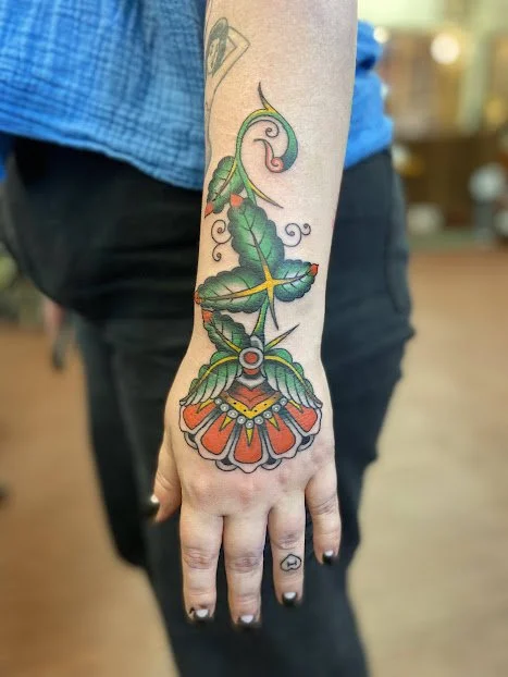 Colorful tattoo of a flower with green leaves and orange petals on a person's hand and forearm.
