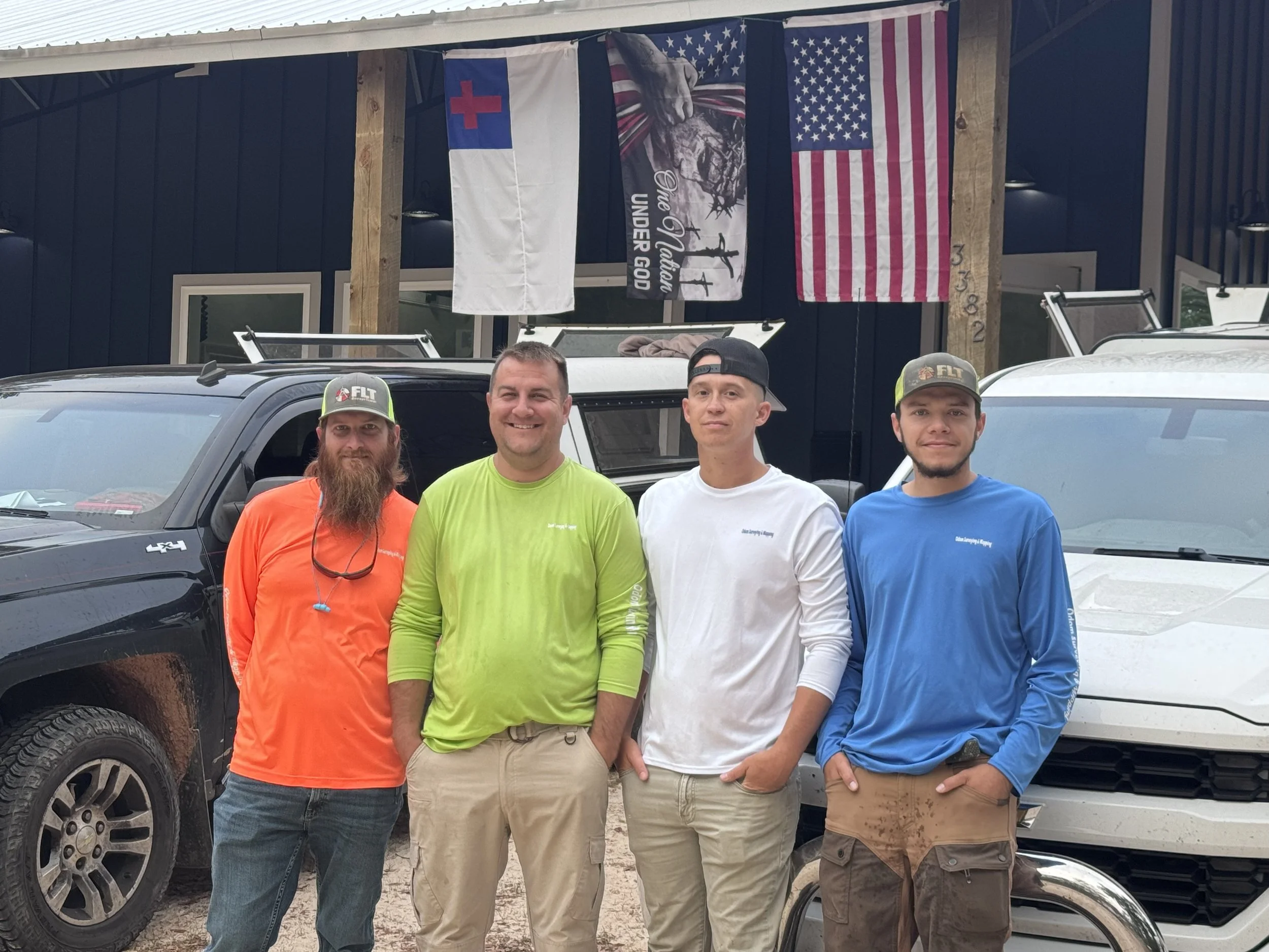 Four young men standing outdoors in front of a black pickup truck, with a building behind them displaying American, Christian, and historical flags.