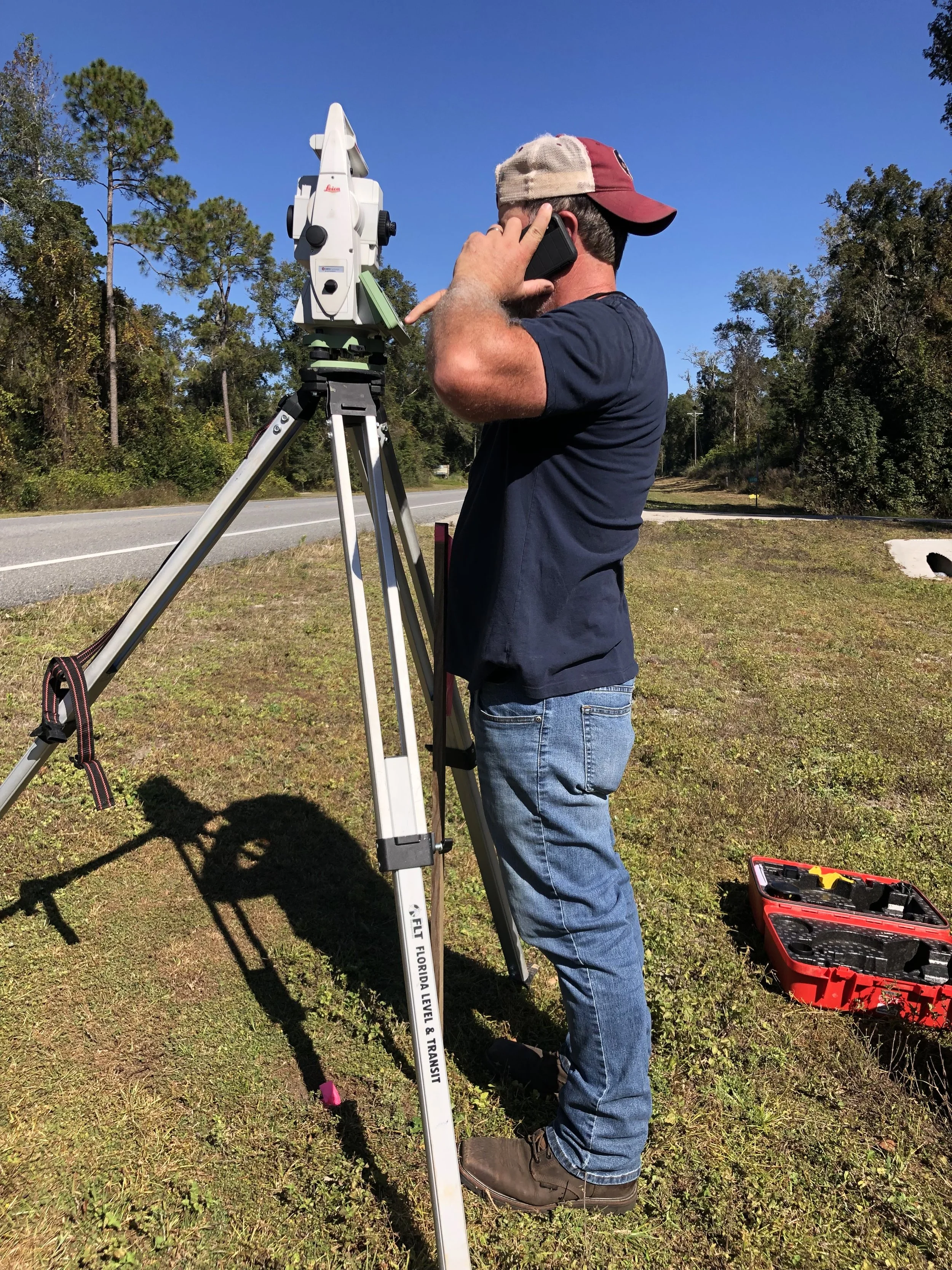 A man wearing a red and beige baseball cap, navy blue shirt, and jeans is standing outdoors on a grassy area, talking on a cellphone while looking through surveying equipment mounted on a tripod. The equipment is a total station used for surveying. There are trees in the background, and a toolbox is on the ground nearby.