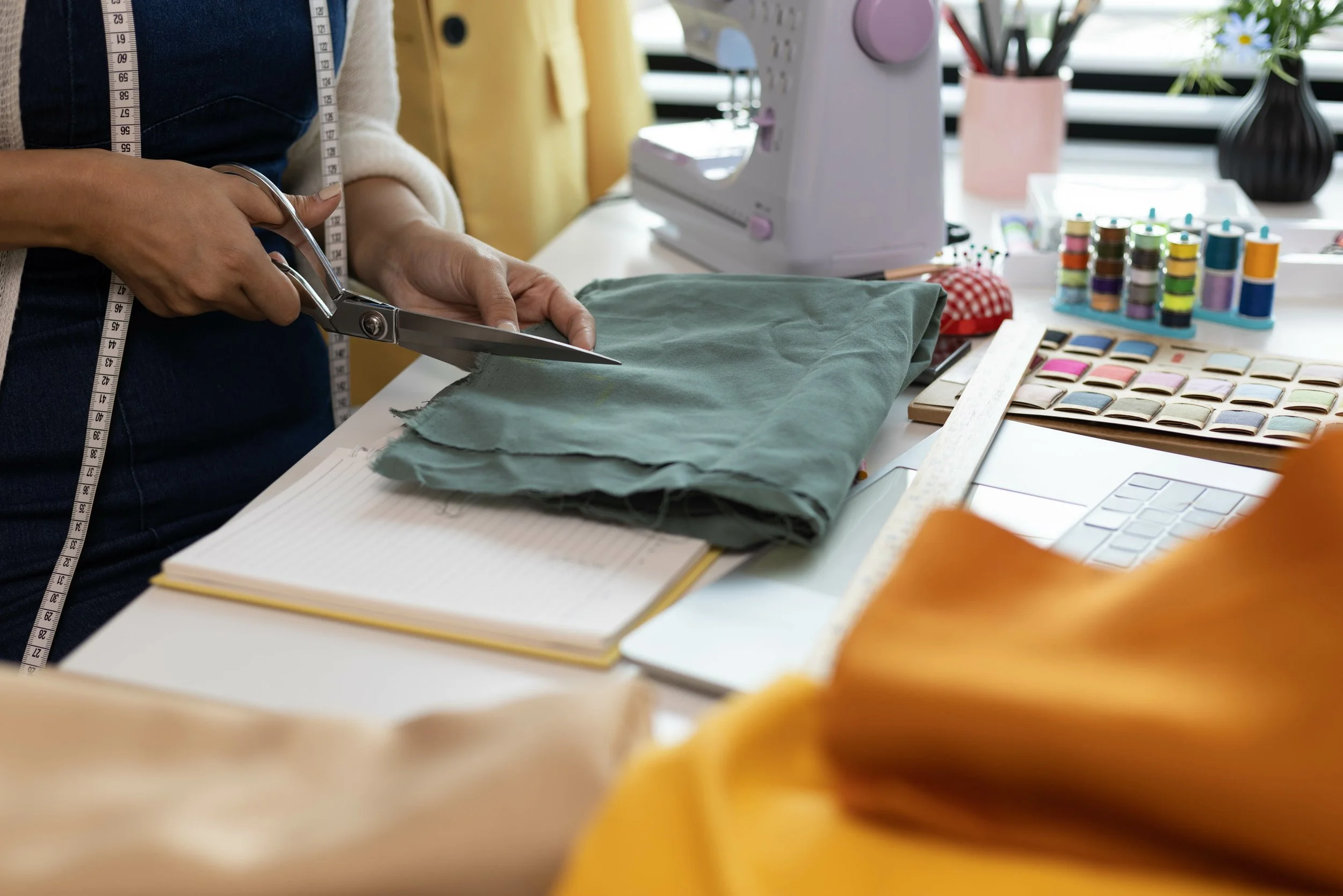 Person cutting fabric on a table with sewing supplies, a measuring tape around their neck, a sewing machine, and color swatches nearby.