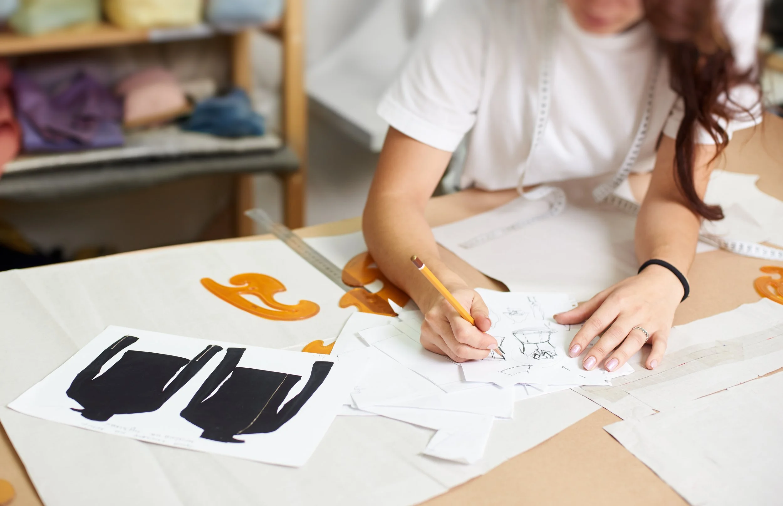 A person working on fashion sketches at a table with fabric swatches, design drawings, and pattern pieces.