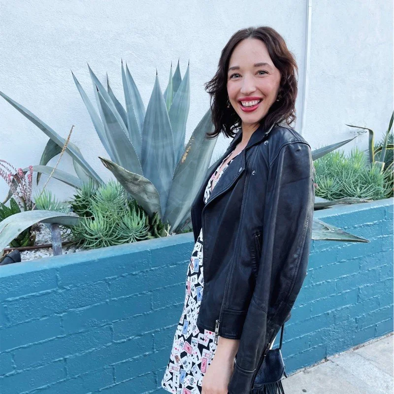 A smiling woman with shoulder-length wavy brown hair wearing a black leather jacket over a patterned dress, standing outdoors beside a blue brick wall with large agave and succulent plants.