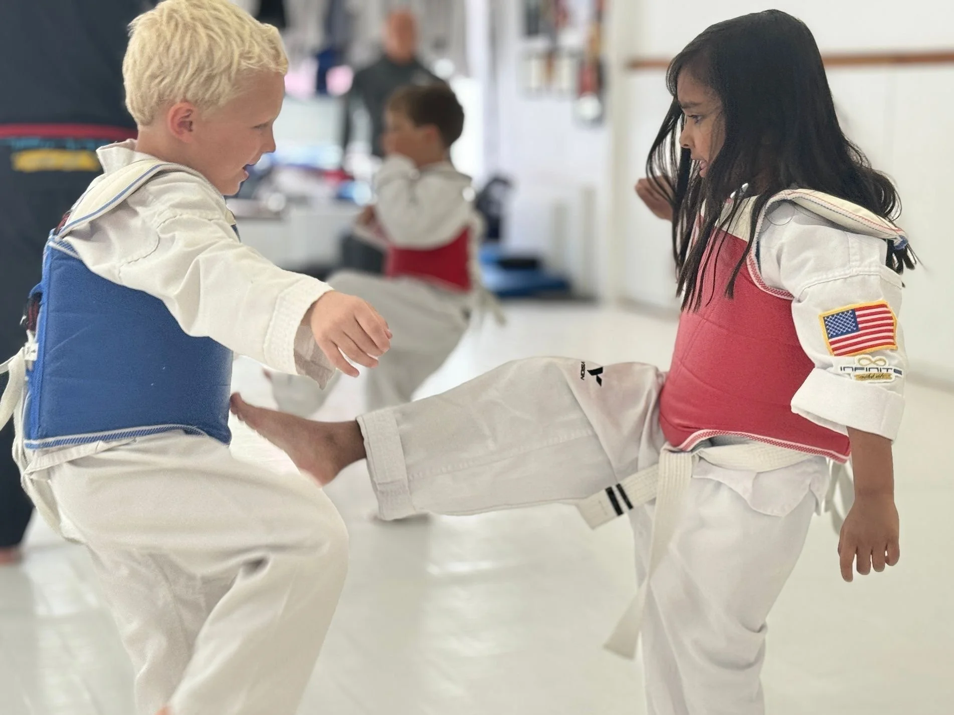 Two children practicing martial arts in a dojo, with one girl blocking a kick from a boy. Both are wearing martial arts uniforms with patches, and other children and an instructor are in the background.