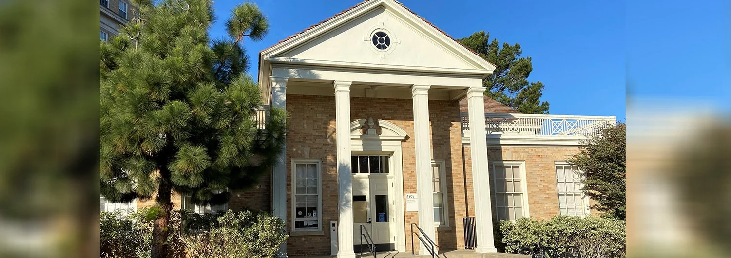 Front view of a brick building with white columns and a pediment, surrounded by greenery and trees, under a clear blue sky.
