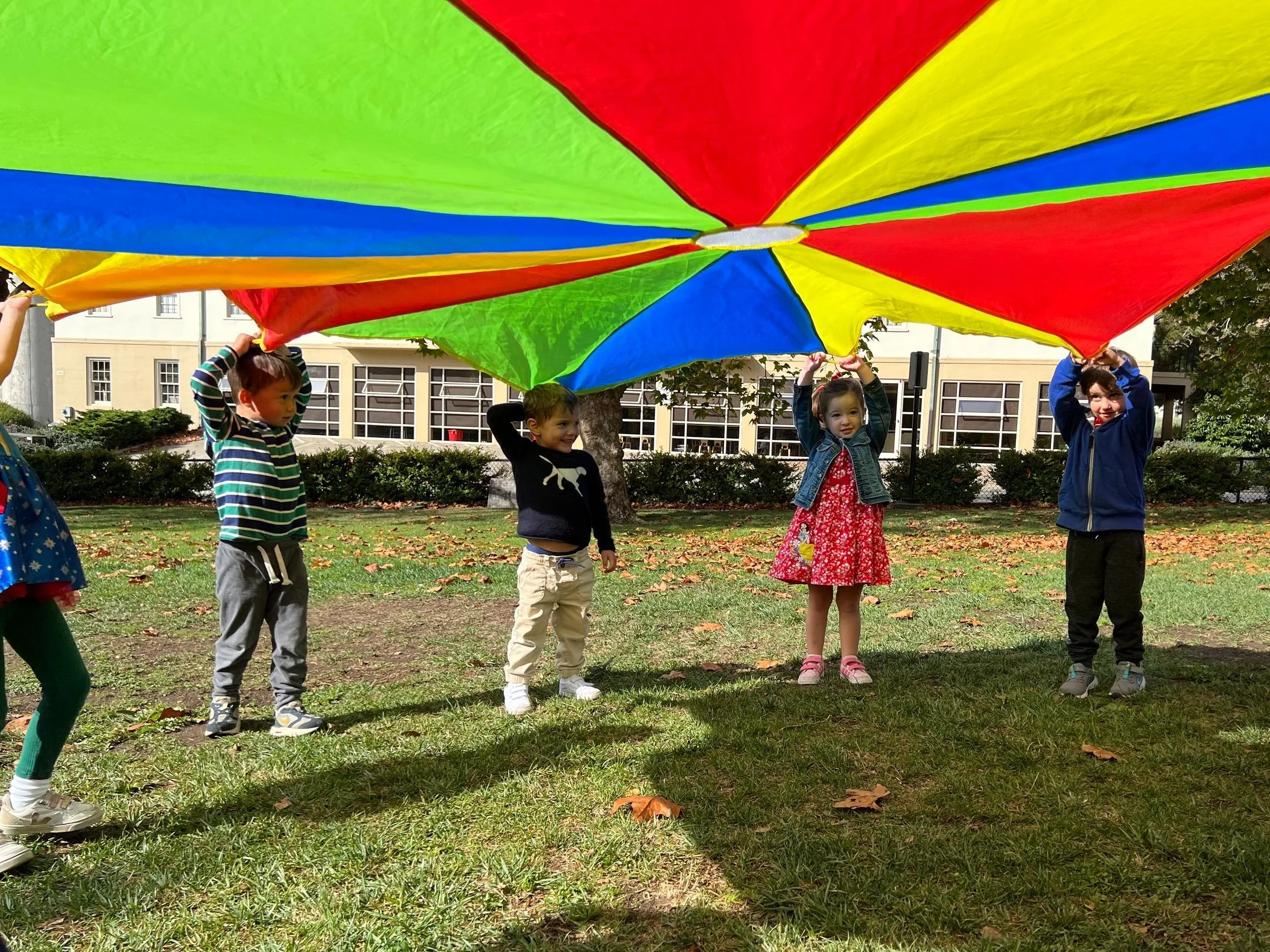 Four children playing outdoors with a colorful parachute, standing in a park with fallen leaves and a building in the background.