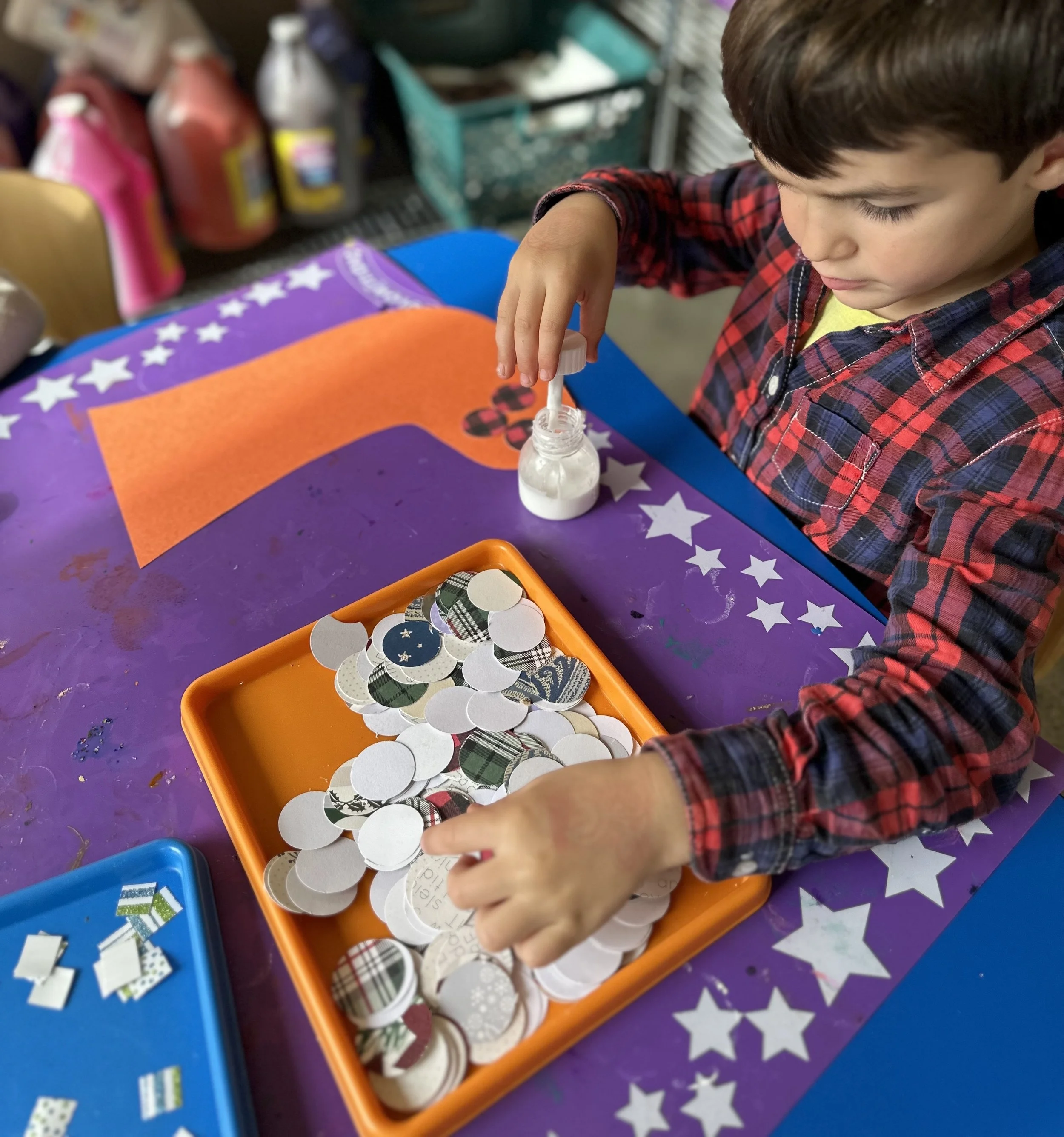 A young boy in a red plaid shirt working with holiday-themed paper circles and glue at a craft table.