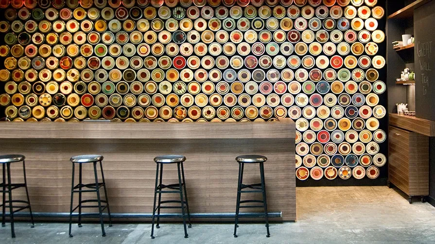 A colorful wall of circular cans or jars behind a wooden bar with four black stools in front, in what appears to be a cafe or coffee shop.