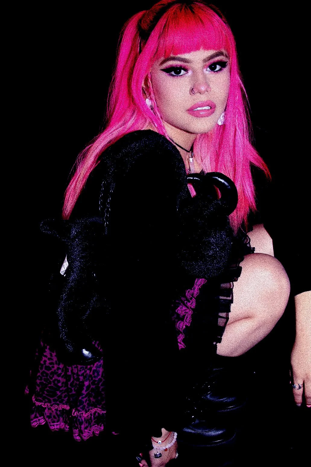 A woman with bright pink hair wearing dark fashion, including a black outfit, sitting against a black background.