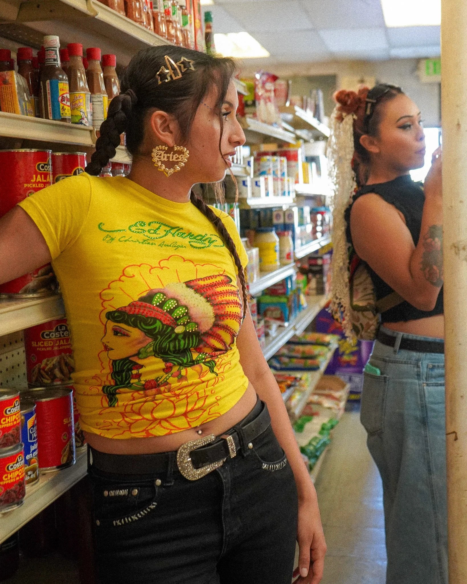 Two women shopping in a grocery store aisle, one wearing a bright yellow T-shirt with a graphic of a woman in historic attire, black jeans, and accessories, and the other in a black crop top and jeans, with shelves of canned goods and condiments behind them.