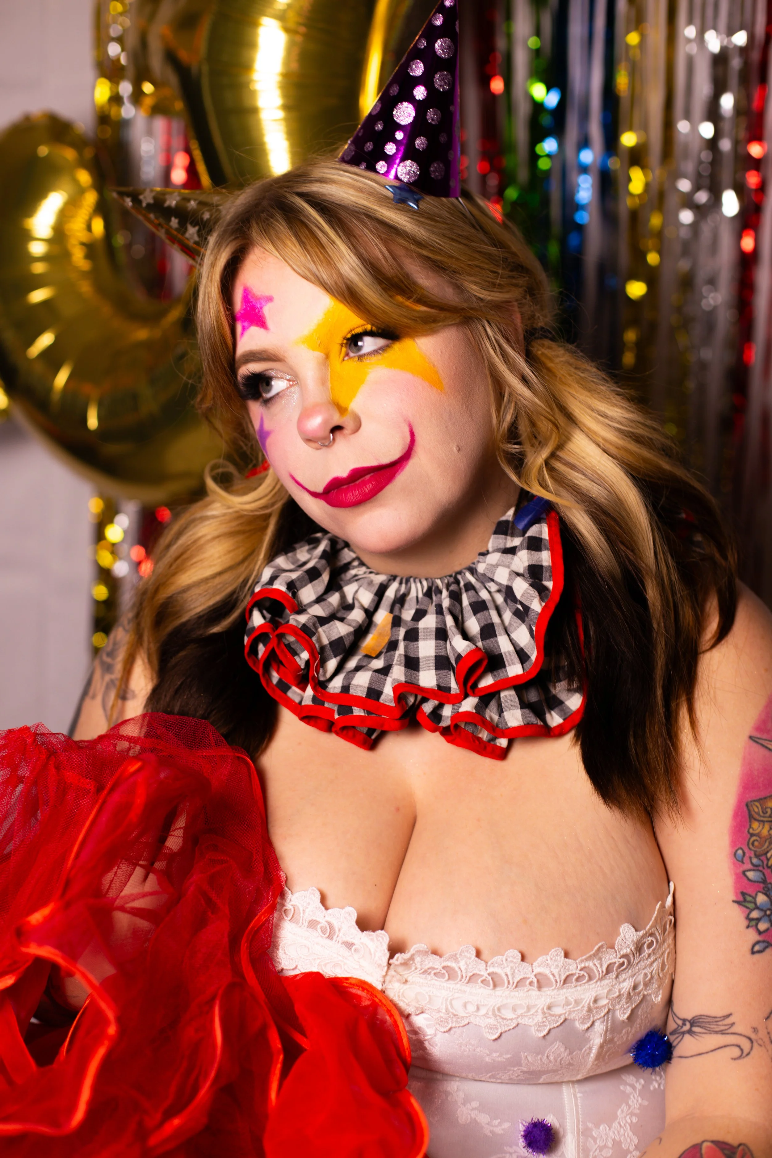 A woman dressed as a clown with colorful face paint, wearing a checkered ruffled collar and a party hat, celebrating at a colorful party.