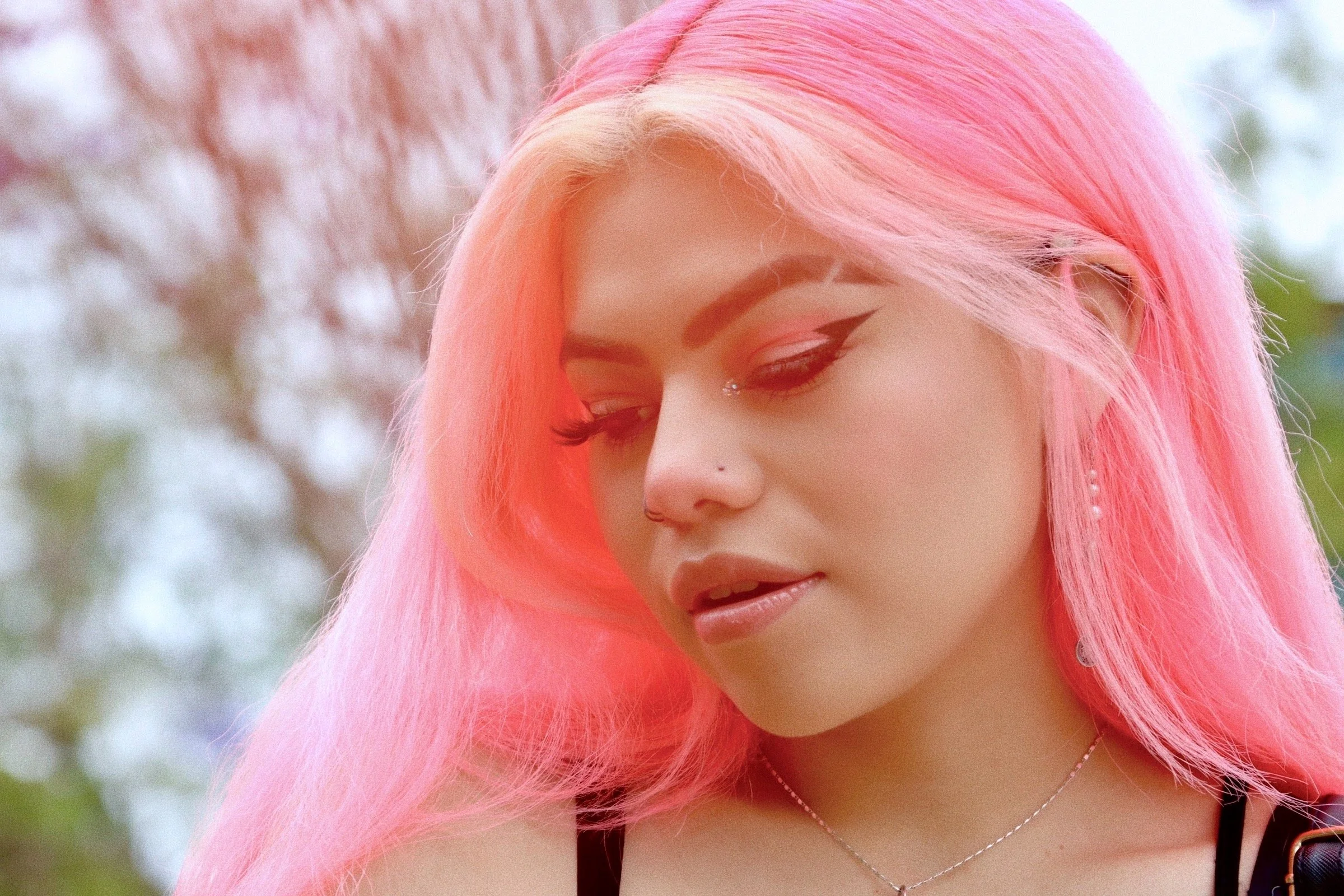 A woman with long, pink hair styled with soft waves, wearing winged eyeliner, long eyelashes, a nose piercing, and a necklace, looking down outdoors with trees in the blurred background.