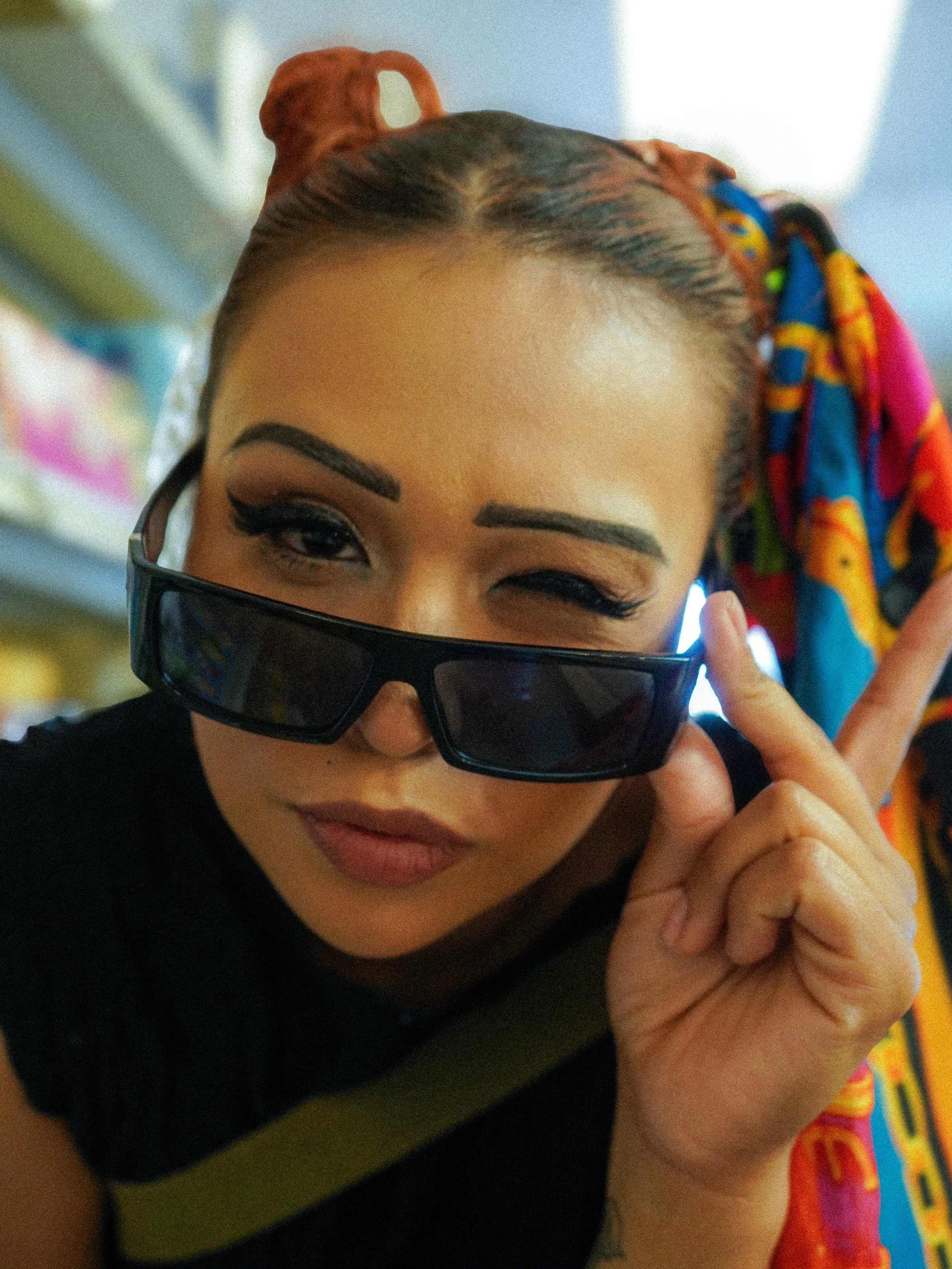 A woman with dark hair styled in braids, wearing a colorful headscarf, black sunglasses, and makeup with winged eyeliner, winking at the camera.
