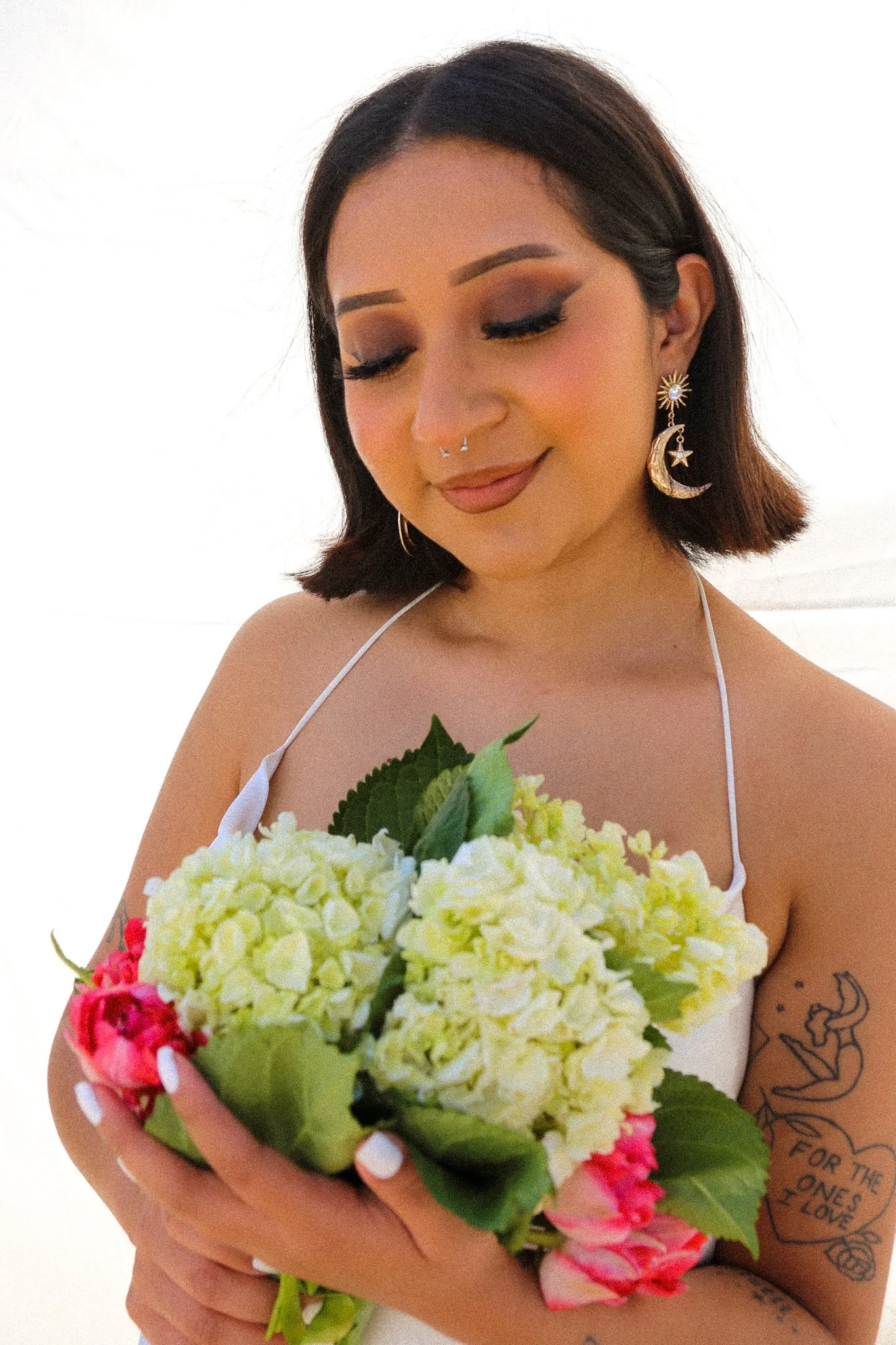 Young woman with dark hair, eye makeup, and moon and star earrings holding a bouquet of white and pink flowers with tattoos on her arm, smiling softly.