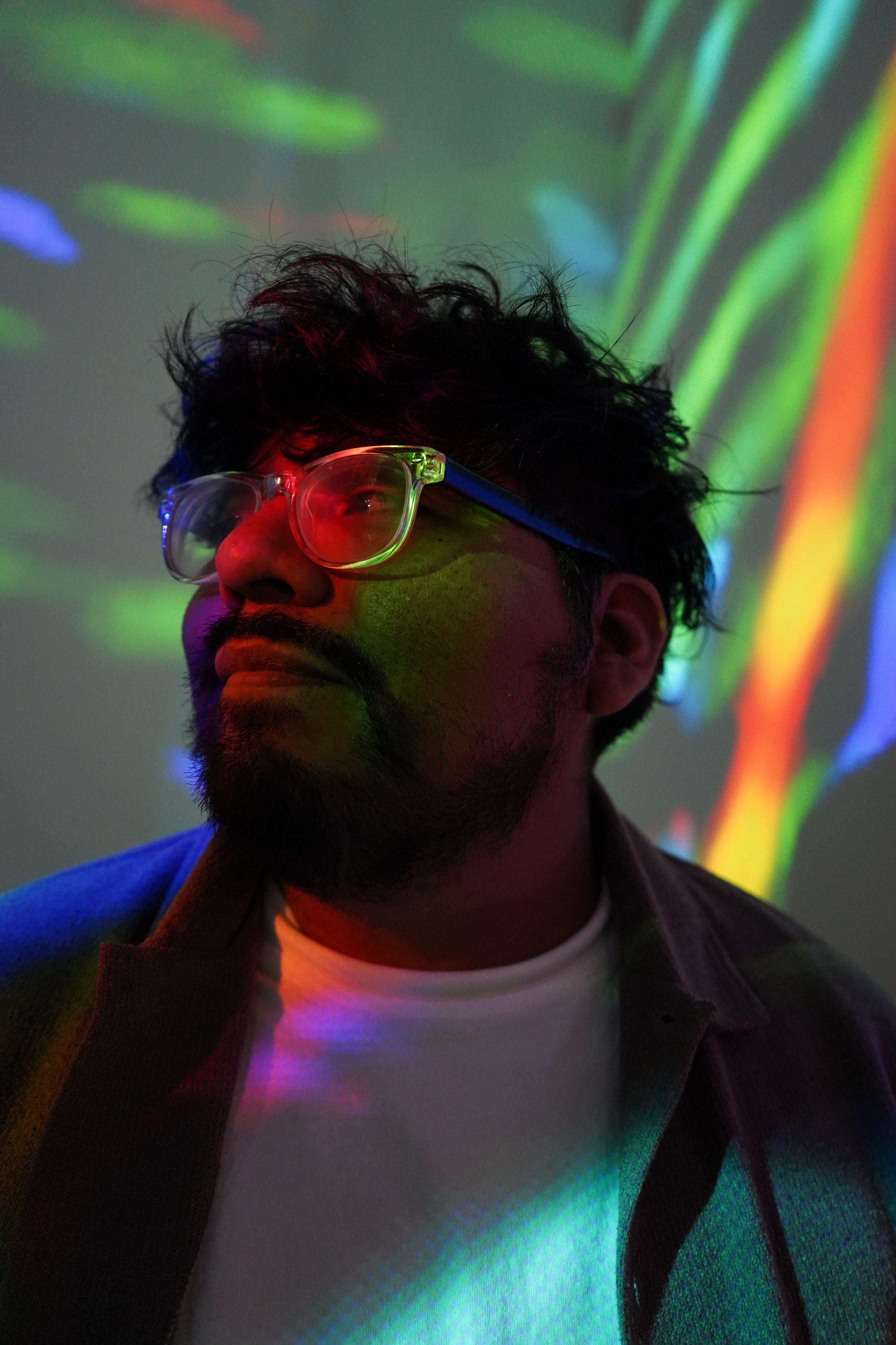 Portrait of a man with glasses, curly hair, and a beard, illuminated by colorful lights and abstract light patterns on the wall behind him.