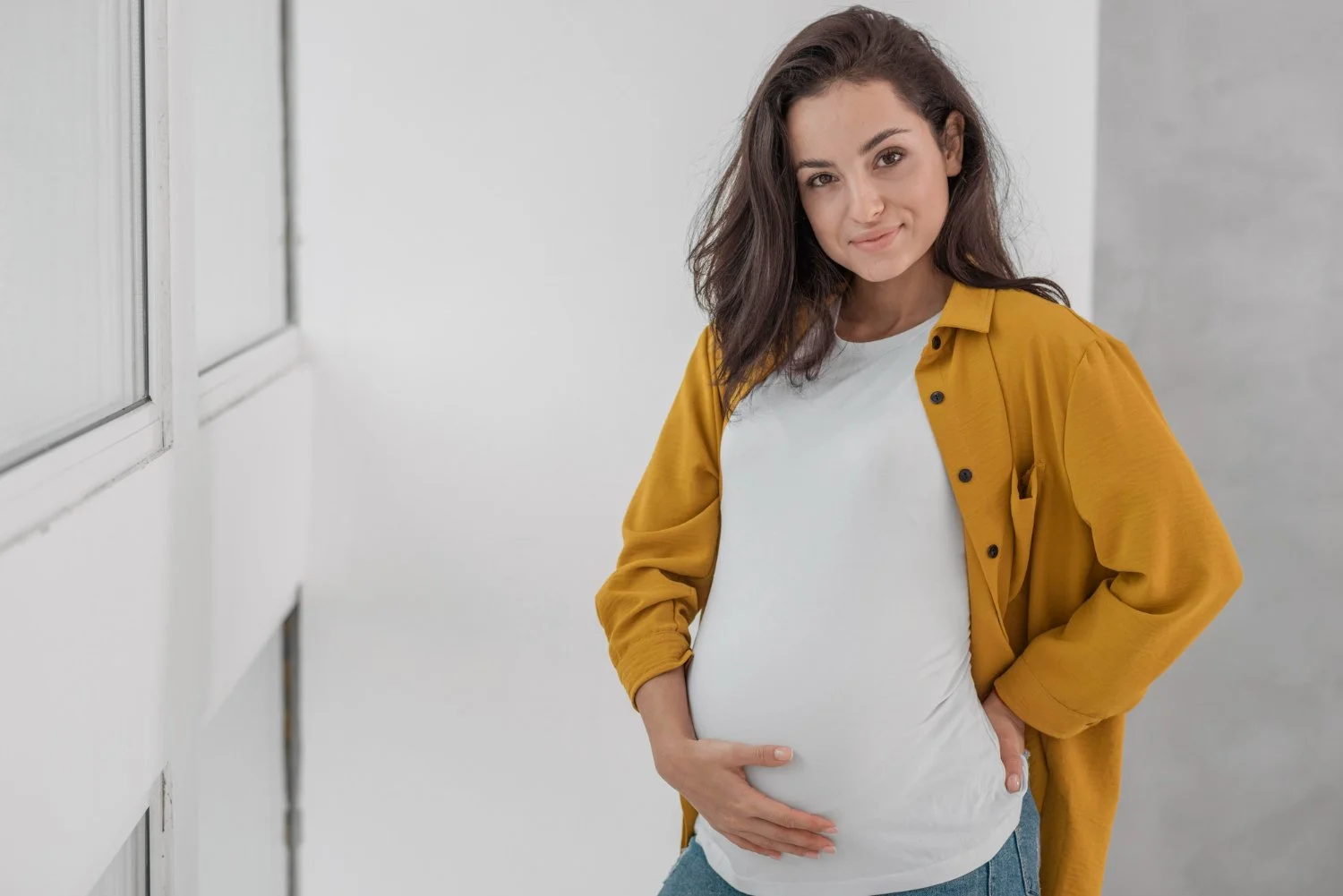 Pregnant woman in white t-shirt and yellow jacket holding her belly and smiling indoors.
