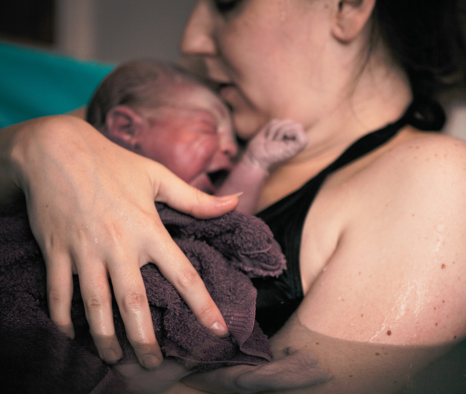 Close-up of a woman holding a newborn baby close to her chest during childbirth, with visible afterbirth marks on her shoulder.