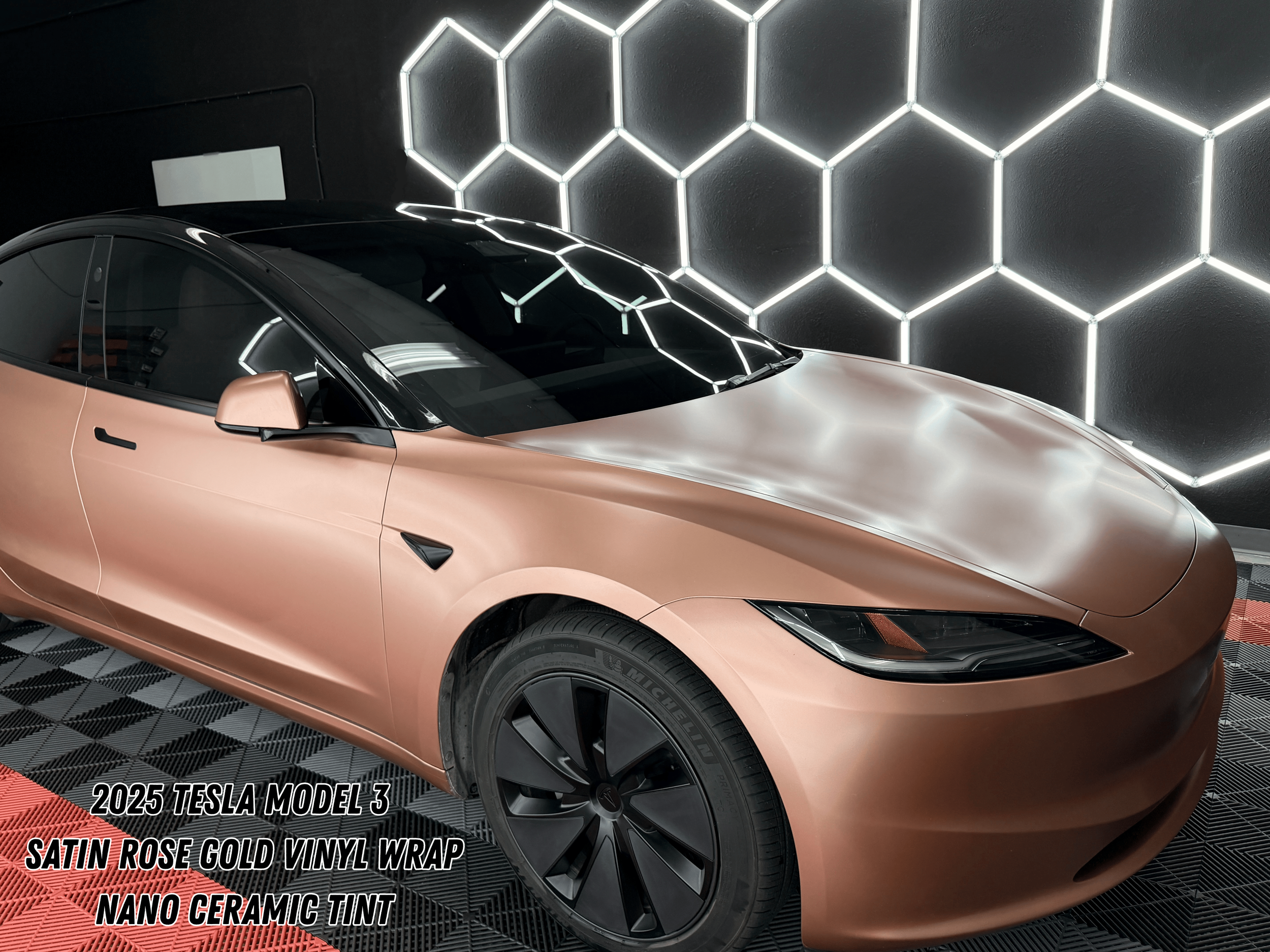 A 2025 Tesla Model 3 vinyl wrapped in satin rose gold vinyl wrap with nano ceramic tint, parked indoors with a black wall featuring illuminated hexagonal pattern, and a checkered floor in black and red.