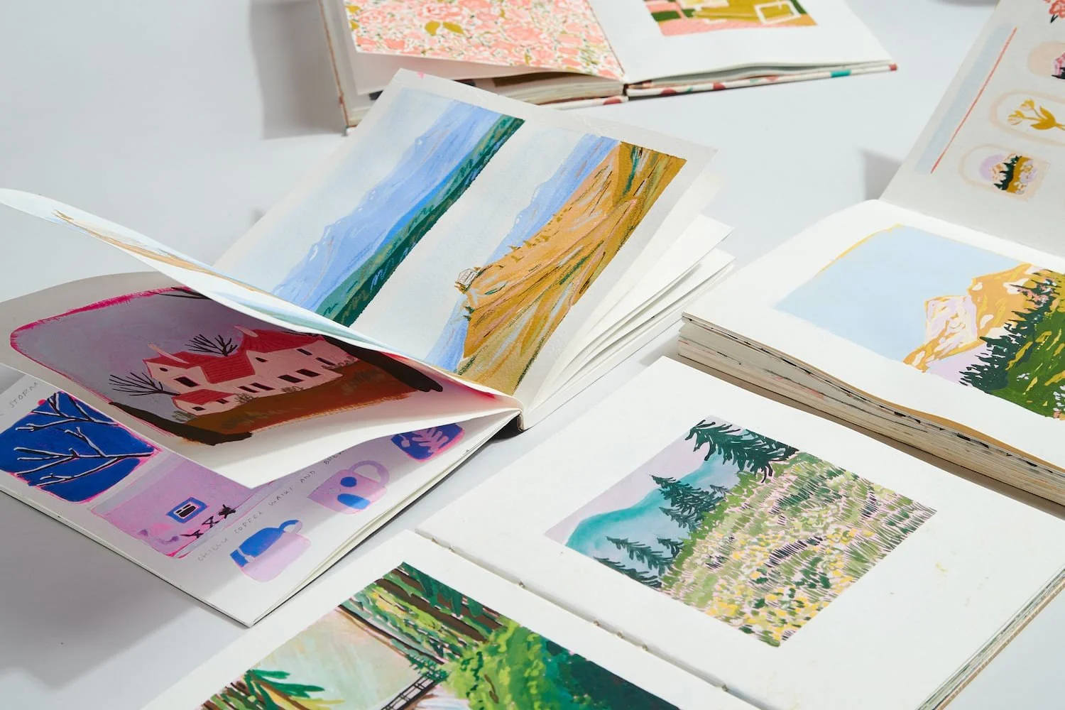 That Creative Space, Open sketchbooks displaying colorful landscape paintings, with scenes of mountains, lakes, trees, and fields spread on a white surface.