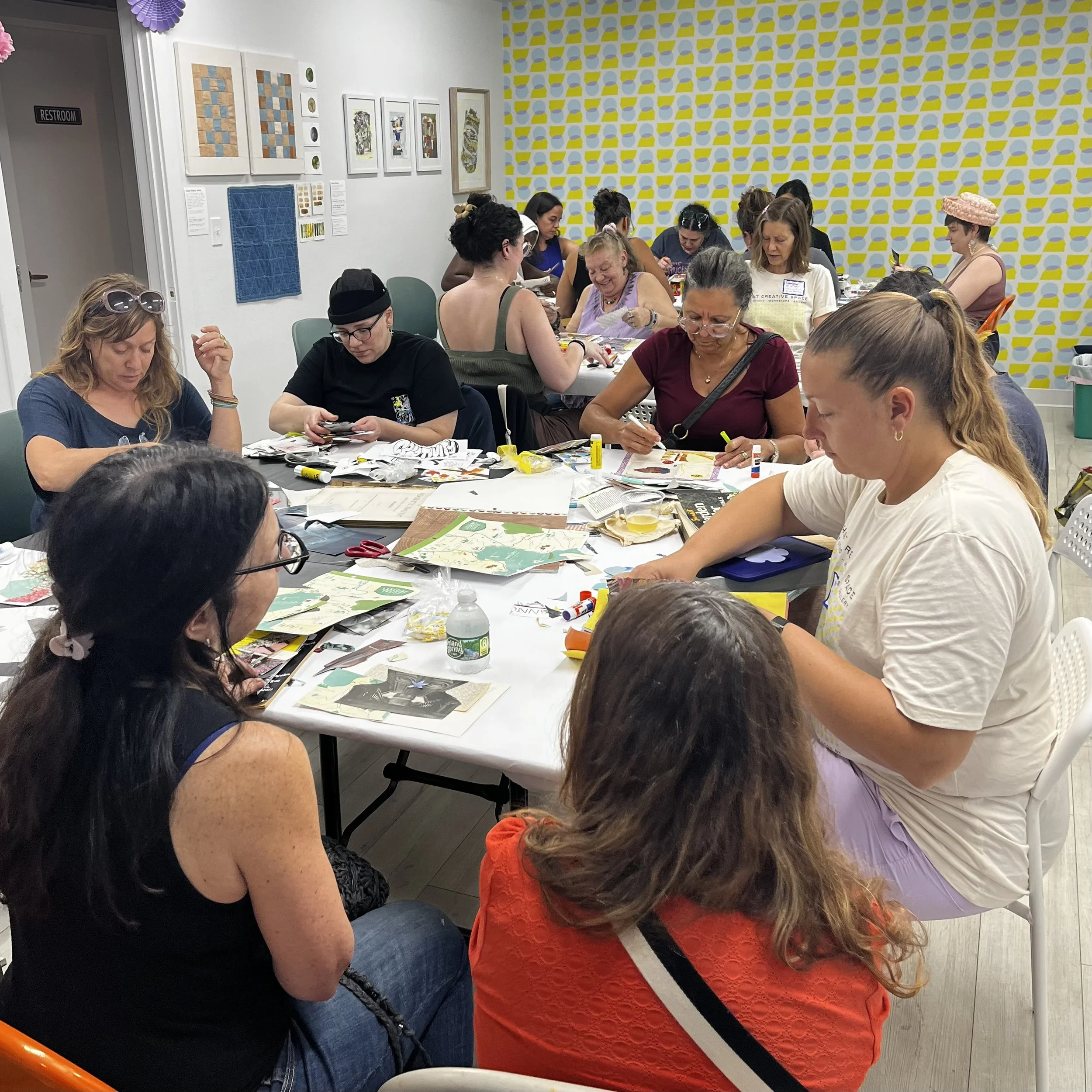 That Creative Space, Open watercolor painting of a landscape with green trees, houses, hills, and a cloudy sky, on a wooden outdoor table with paint supplies and brushes.