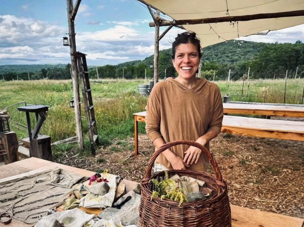 That Creative Space, A smiling woman standing outdoors behind a wooden table with fabric and rocks, holding a wicker basket. The setting appears to be a rural or farm environment with green fields, mountains in the background, and a canopy overhead.