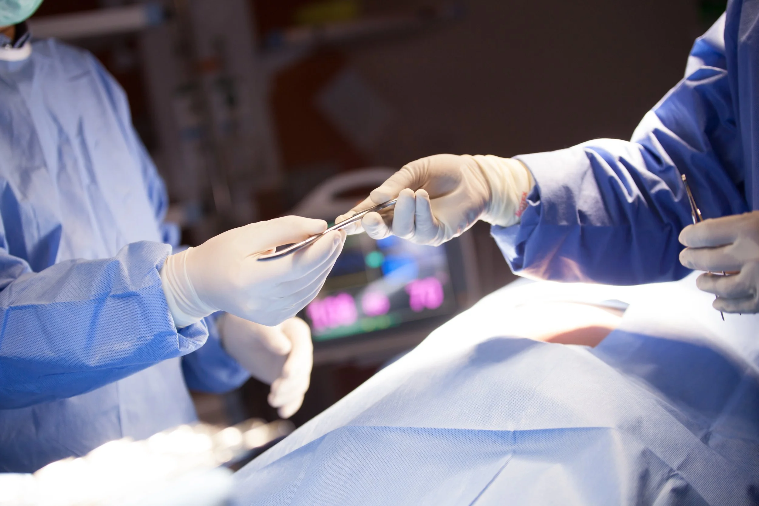 Surgeons in blue gowns and gloves performing a surgical procedure, exchanging surgical instruments over a patient.