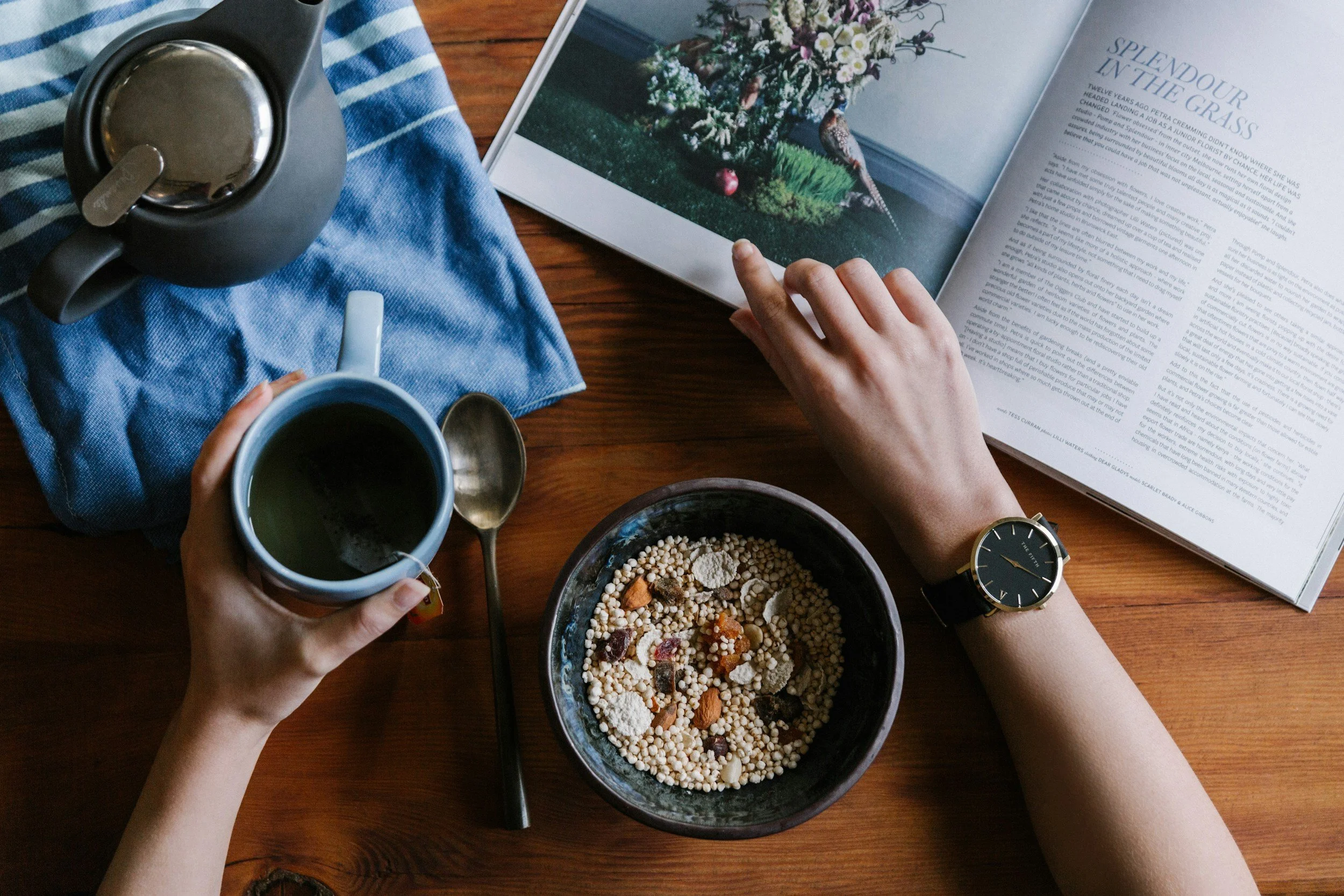 A person holding a cup of tea or coffee, with a bowl of cereal on a wooden table, and an open magazine.,