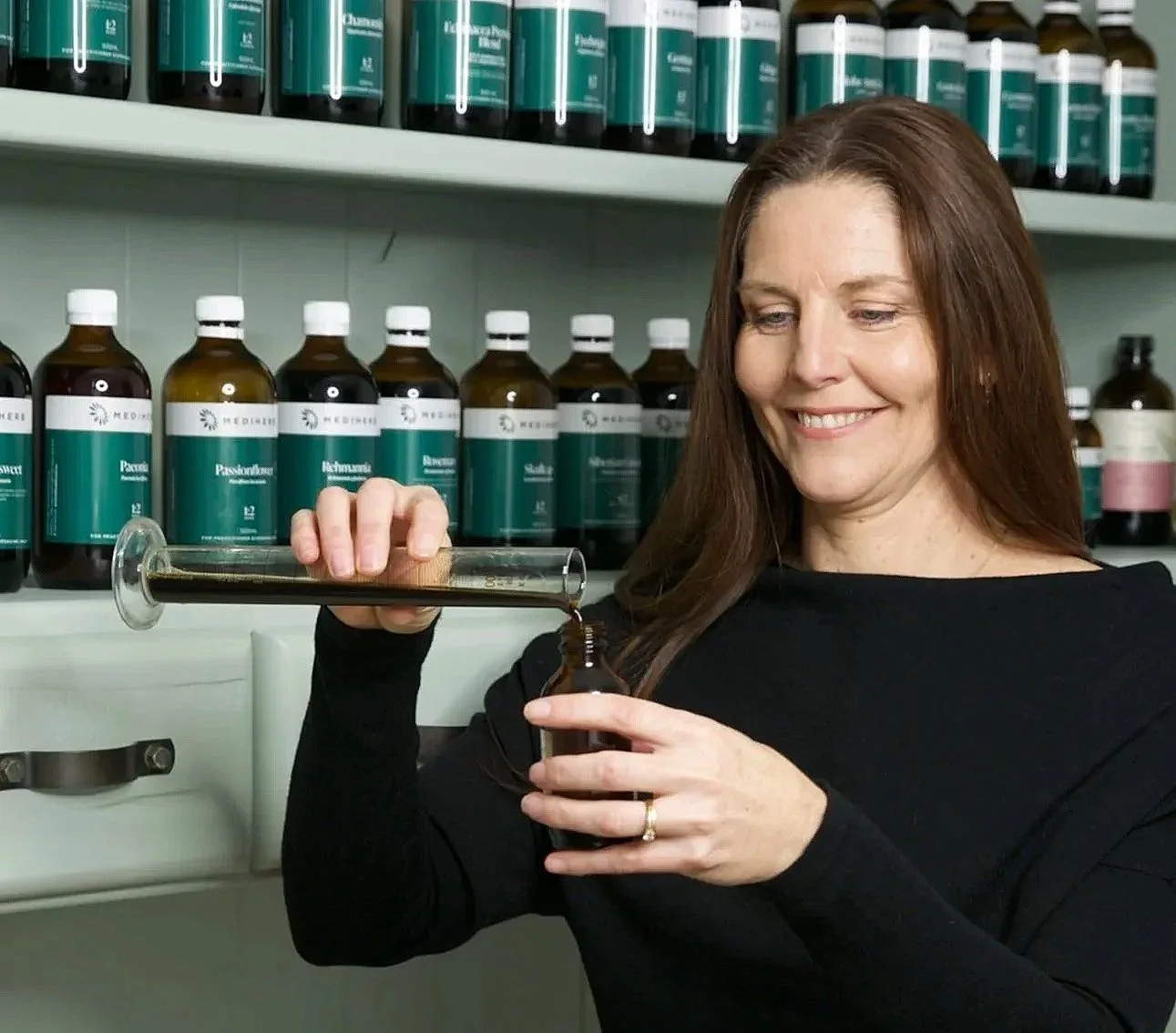 A woman with brown hair smiling at a pharmacy or herbal store, filling a small bottle with liquid from a large syringe, with shelves of medicine bottles in the background.