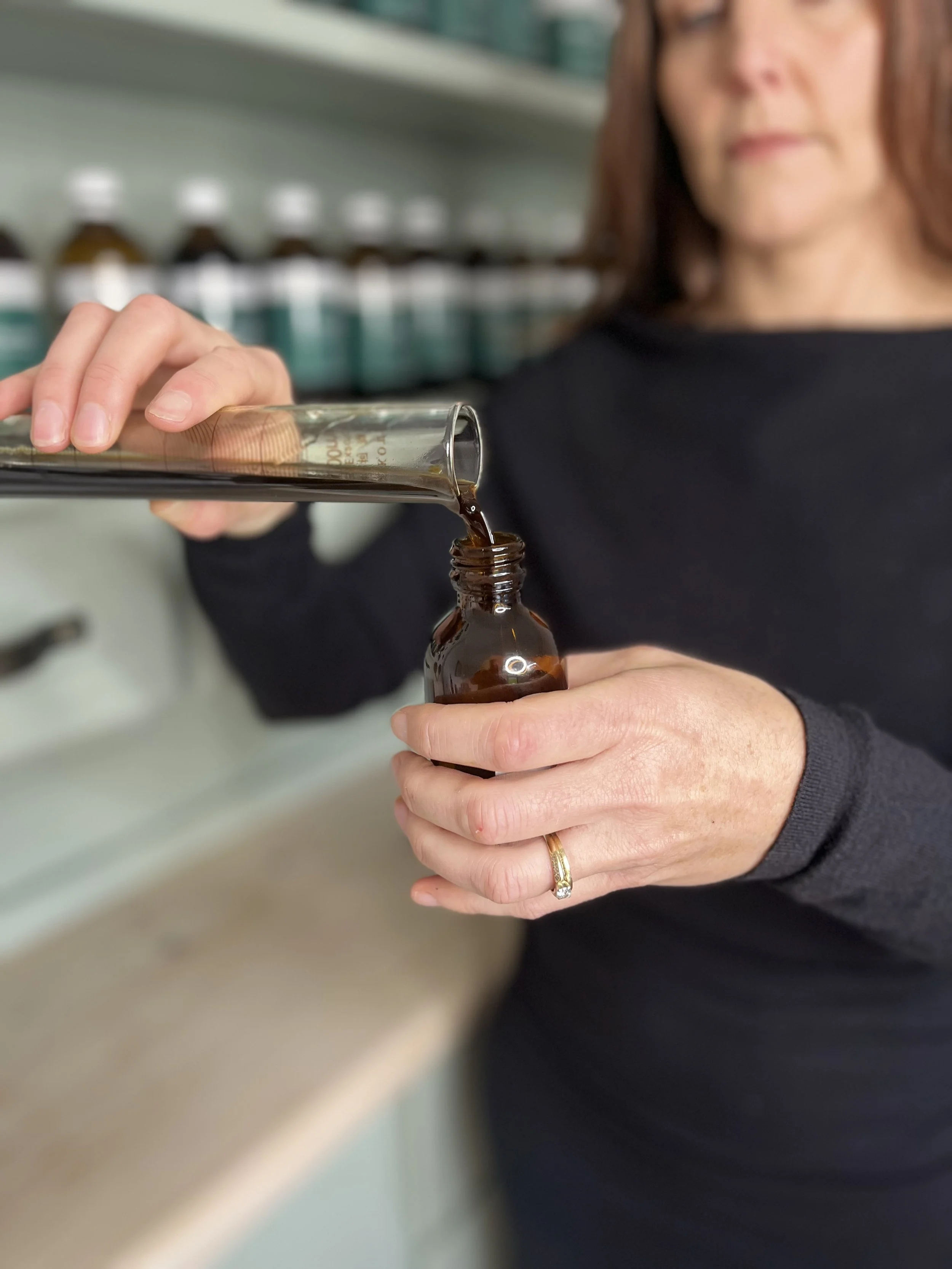 A woman with a gold wedding ring is pouring a dark liquid from a glass bottle into a small brown bottle, standing in front of shelves with small bottles.