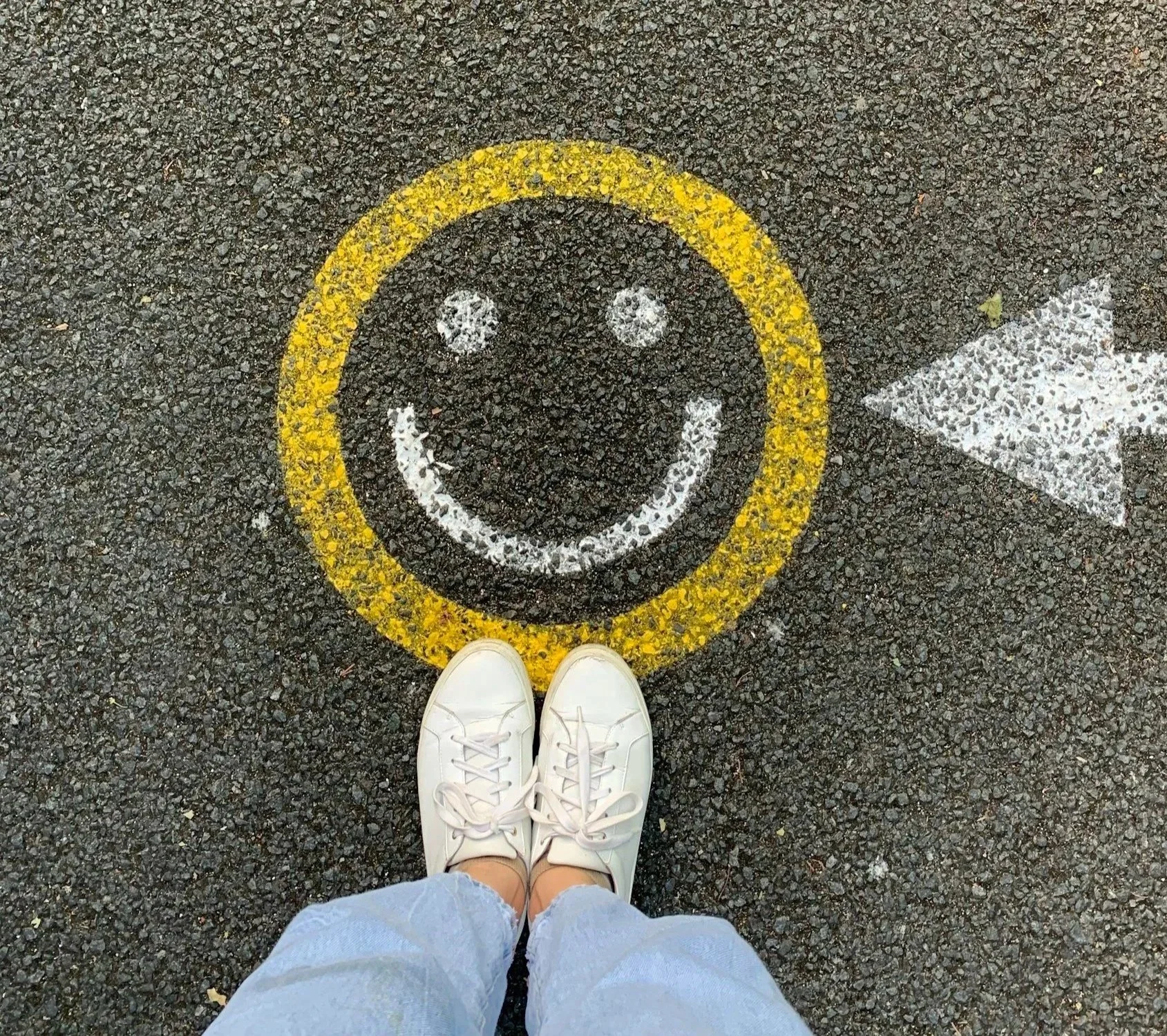 Person standing on sidewalk in front of a smiley face and arrow painted on the ground, wearing white sneakers and light blue jeans.
