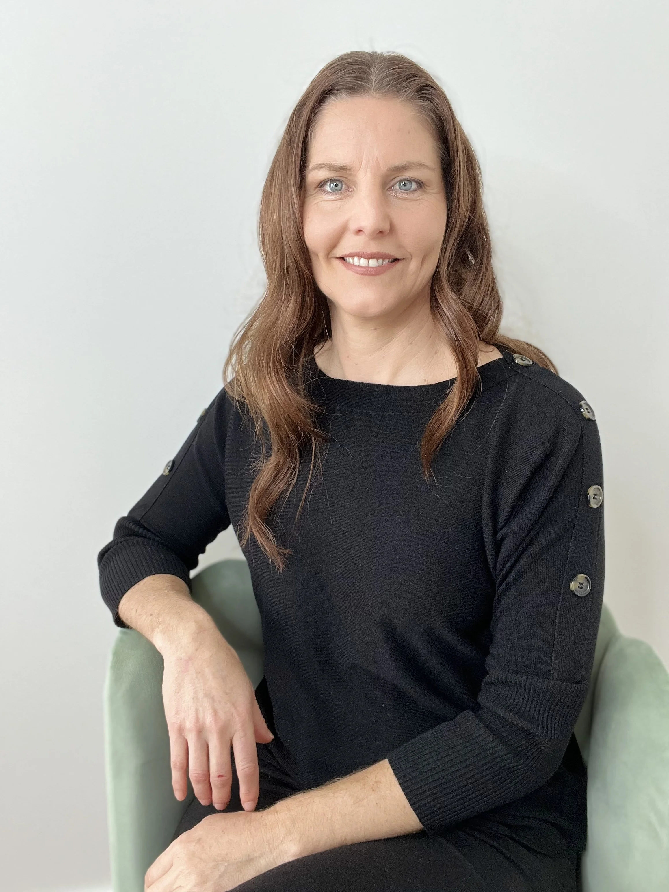 Close-up of a woman with shoulder-length brown hair, light blue eyes, smiling, wearing a black top with buttons on the shoulders, sitting on a green armchair against a plain white wall.