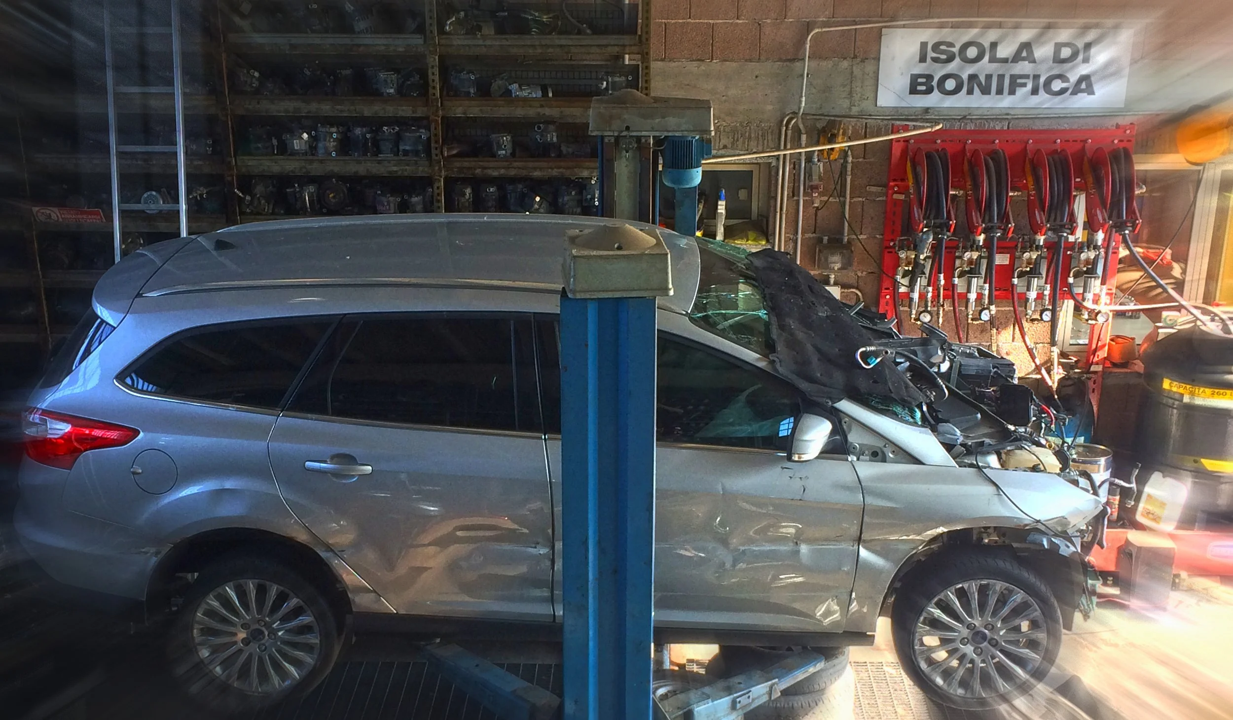 Auto danneggiata con il cofano aperto in un'officina, scaffali con ricambi e attrezzi, cartello con scritto 'Isola di Bonifica'.