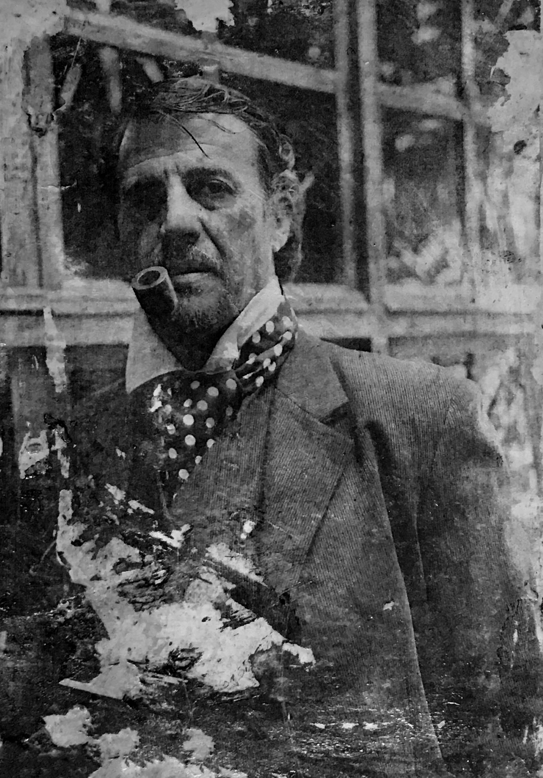 A black and white photograph of a man with a pipe in his mouth, dressed in a suit and polka dot tie, standing outdoors with a brick or stone structure in the background.