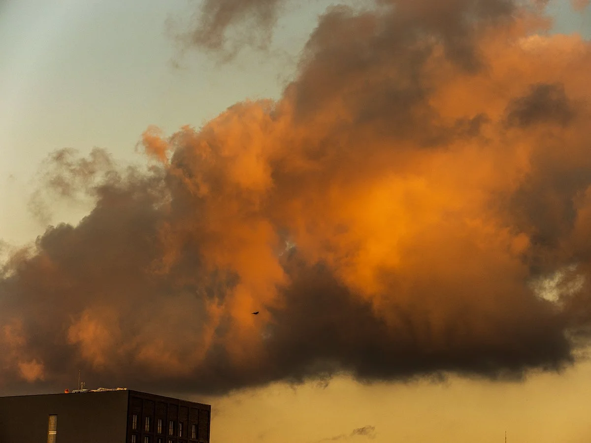 Large orange and dark gray cloud of smoke in the sky above a building at sunset.