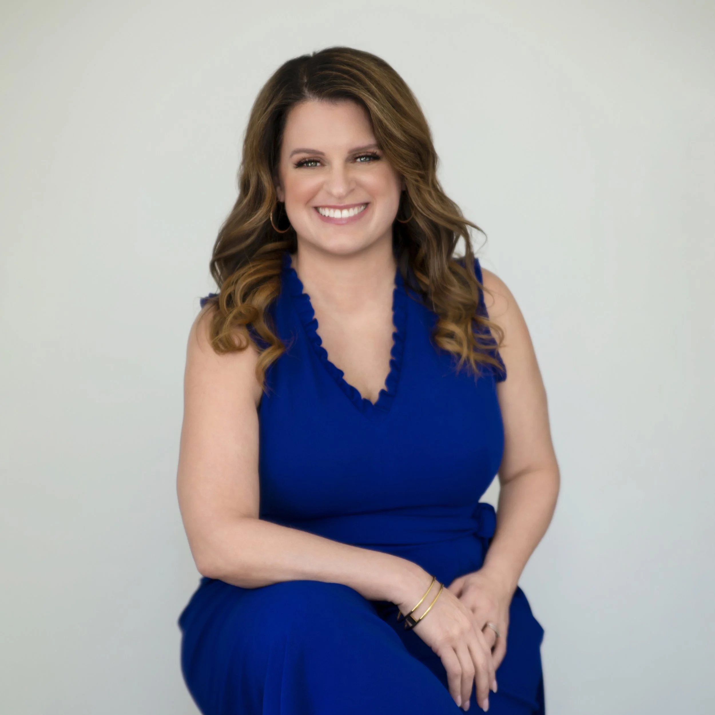 A woman with long, wavy brown hair smiling, wearing a sleeveless blue dress, sitting against a plain light background. She is a licensed insurance broker.