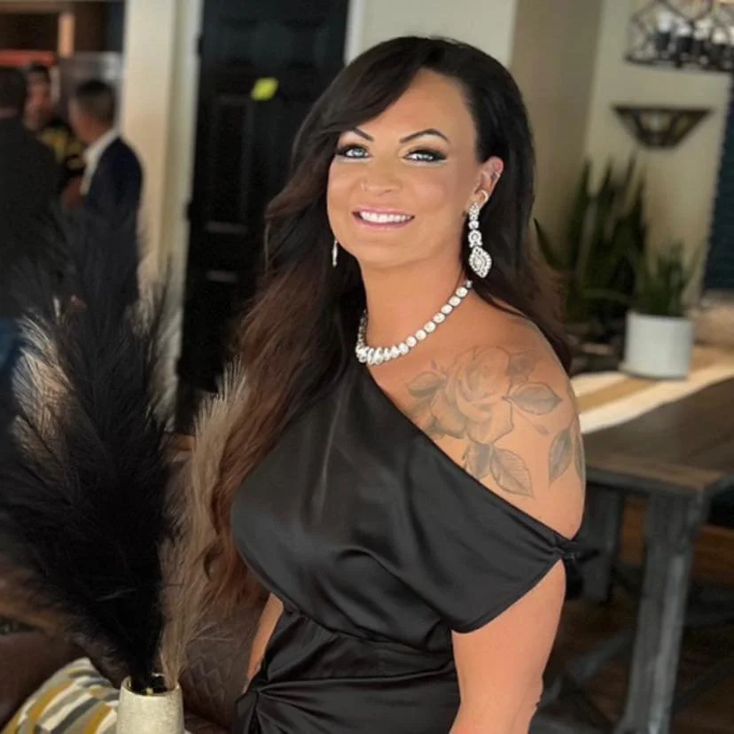 A woman with long dark hair, wearing a black off-shoulder dress, pearl necklace, and matching earrings, smiling inside a decorated room with plants and people in the background.