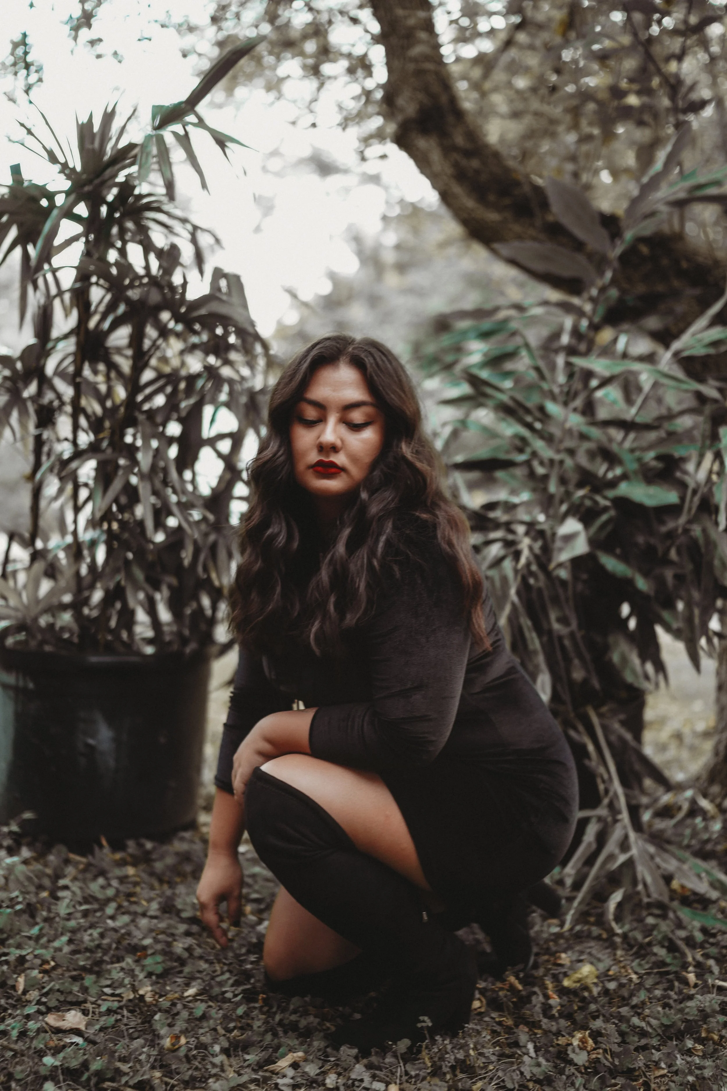 A woman, agency owner Grace Kavanaugh, with dark curly hair and red lipstick crouches outdoors, surrounded by large potted plants and trees.