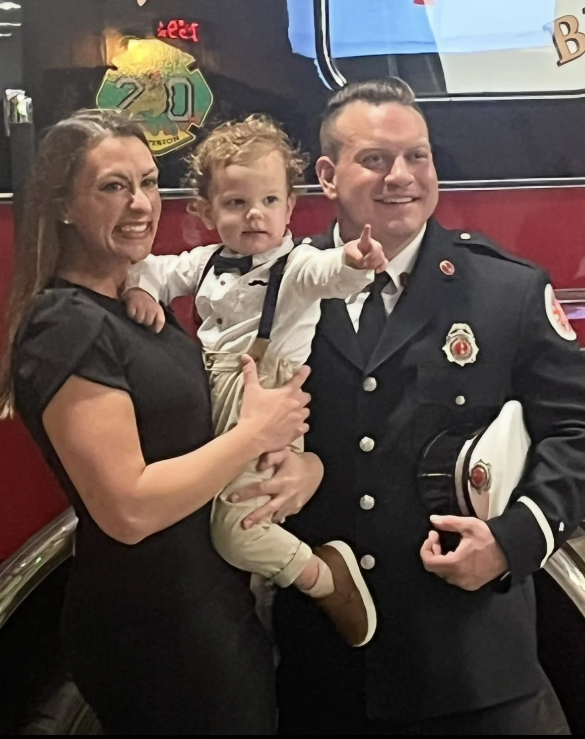 A woman, a young boy, and a man in a firefighter's uniform pose together. The woman is giving a thumbs-up, the boy points into the distance, and the firefighter holds a cap. They are smiling. They Own Ready Set Rescue Training