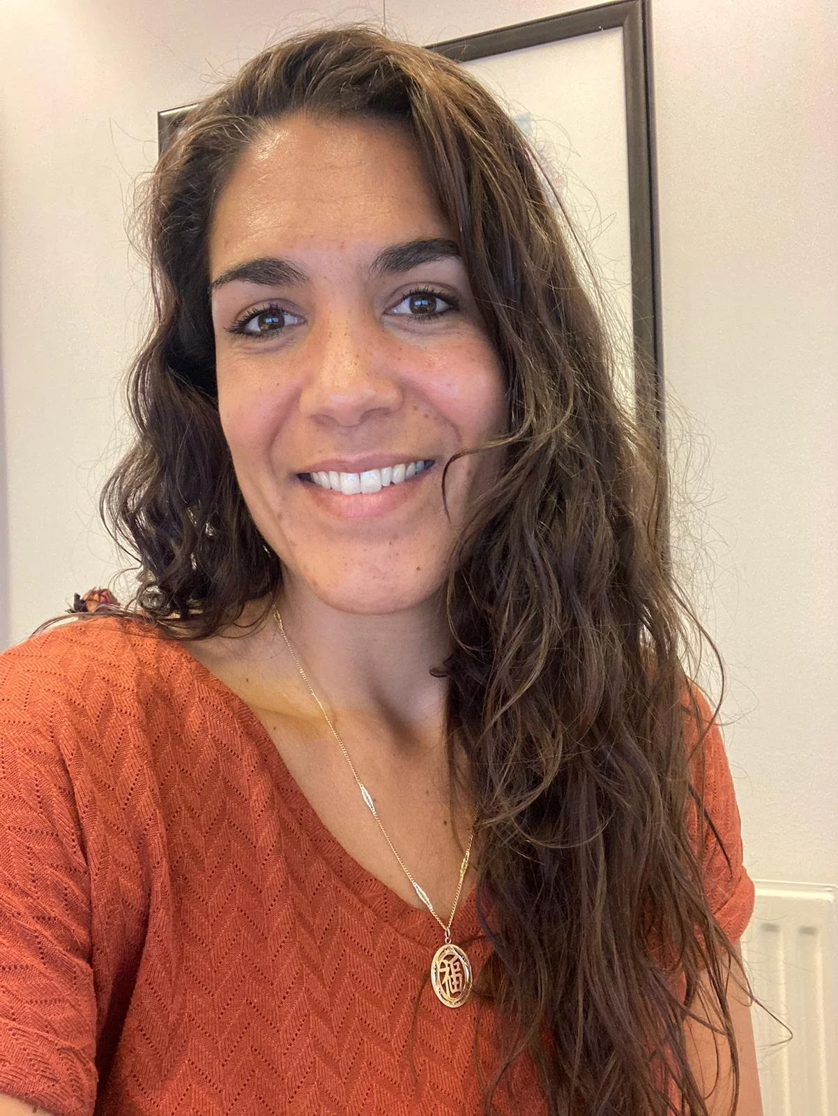 A woman with long, curly brown hair wearing an orange top and a gold necklace, smiling at the camera indoors.