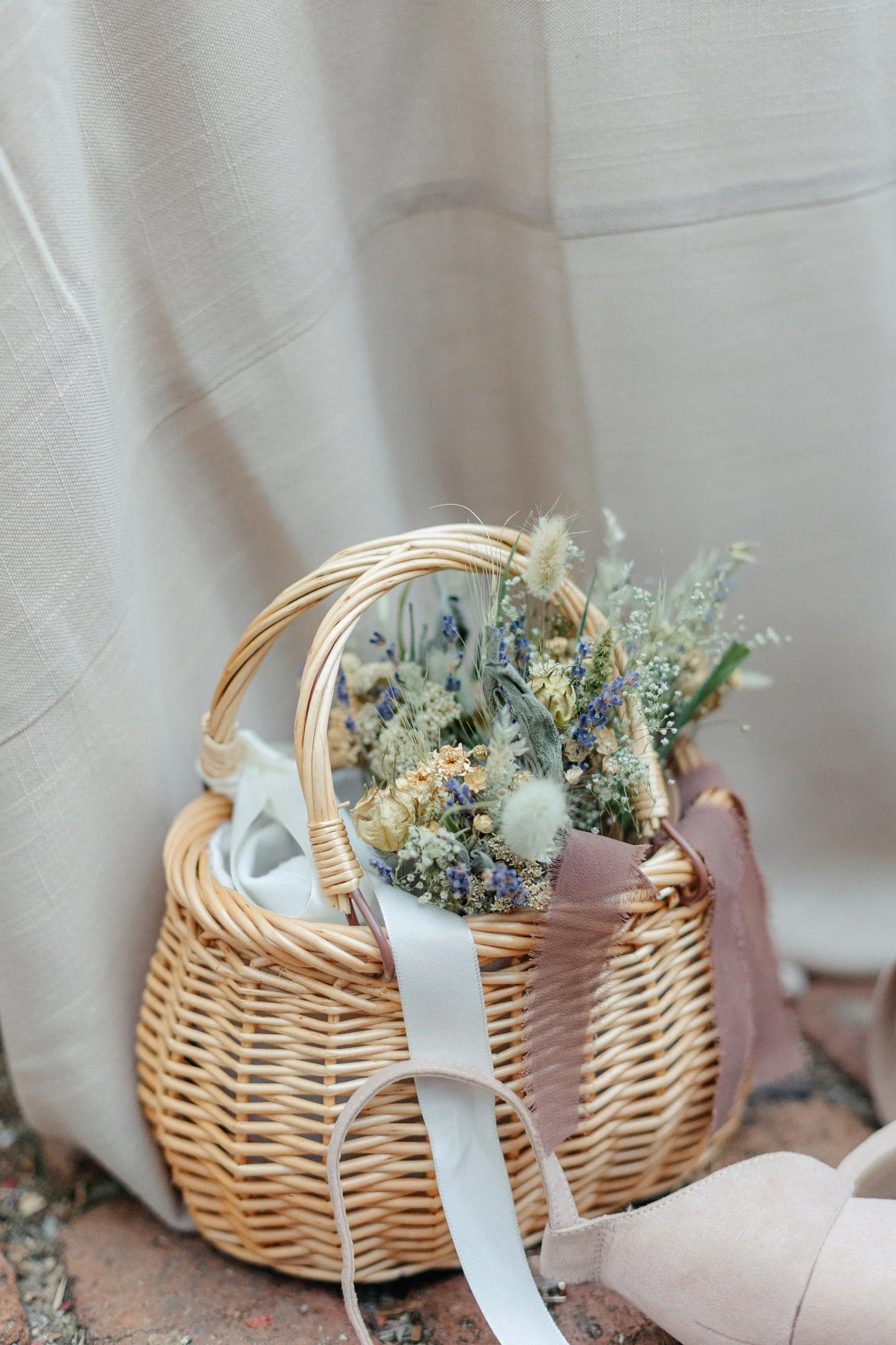 A woven basket filled with dried flowers and ribbons, placed on a brick ground against a beige curtain.