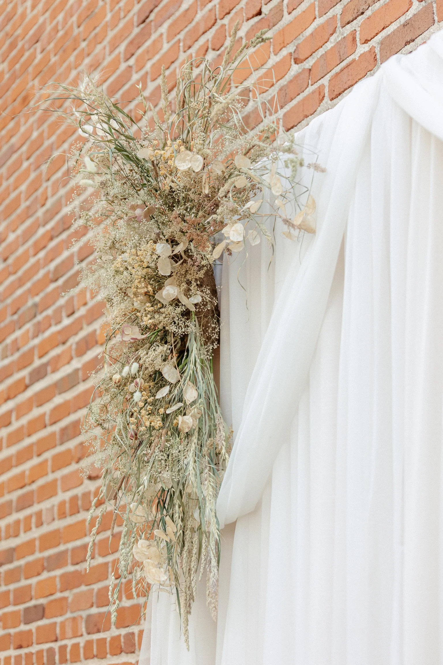A floral arrangement with dried flowers and greenery, attached to a white curtain against a brick wall.