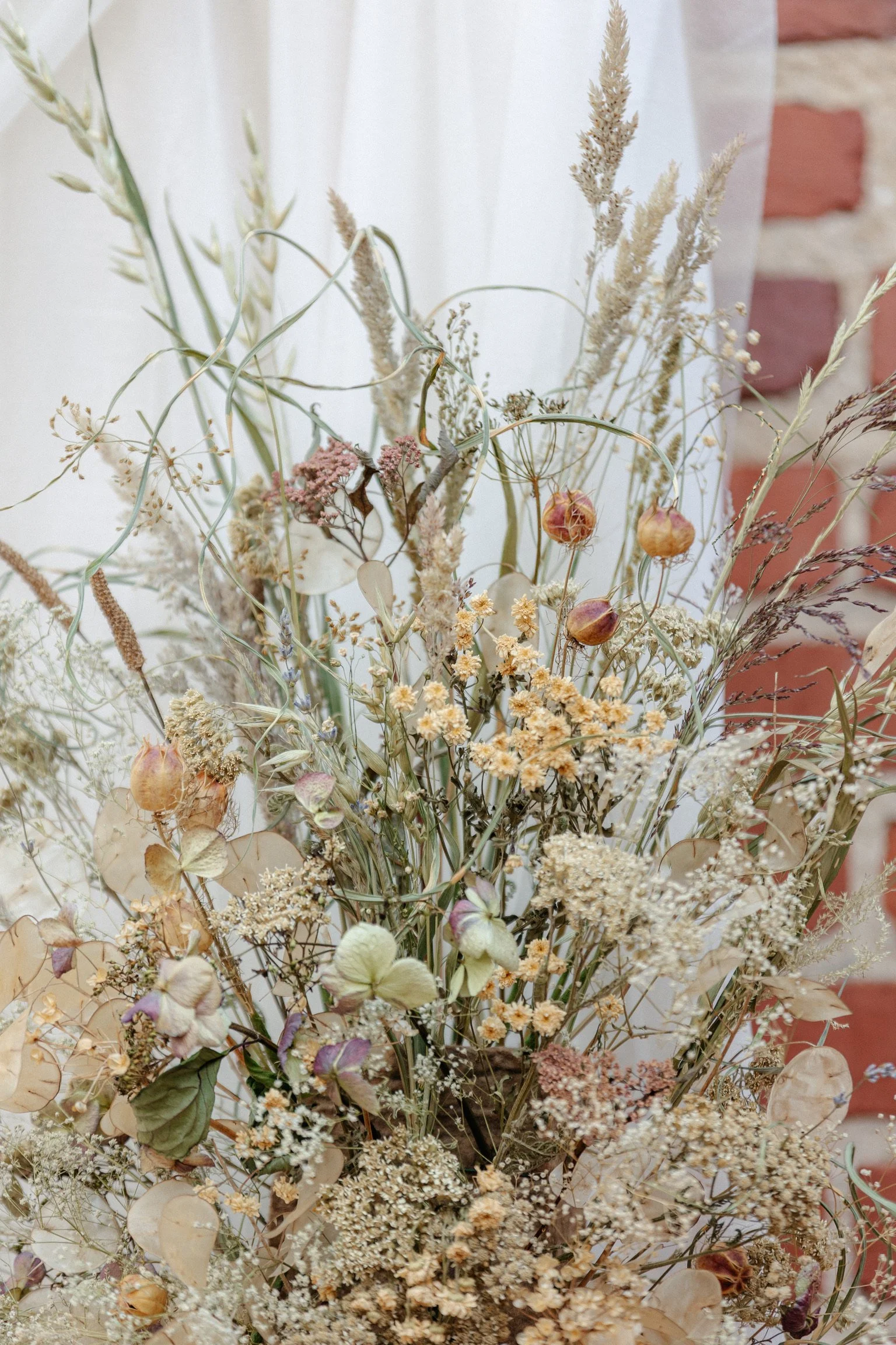 Dried wildflower bouquet with various beige, pink, and purple flowers and grasses, set against a blurred background of a brick wall and white curtain.